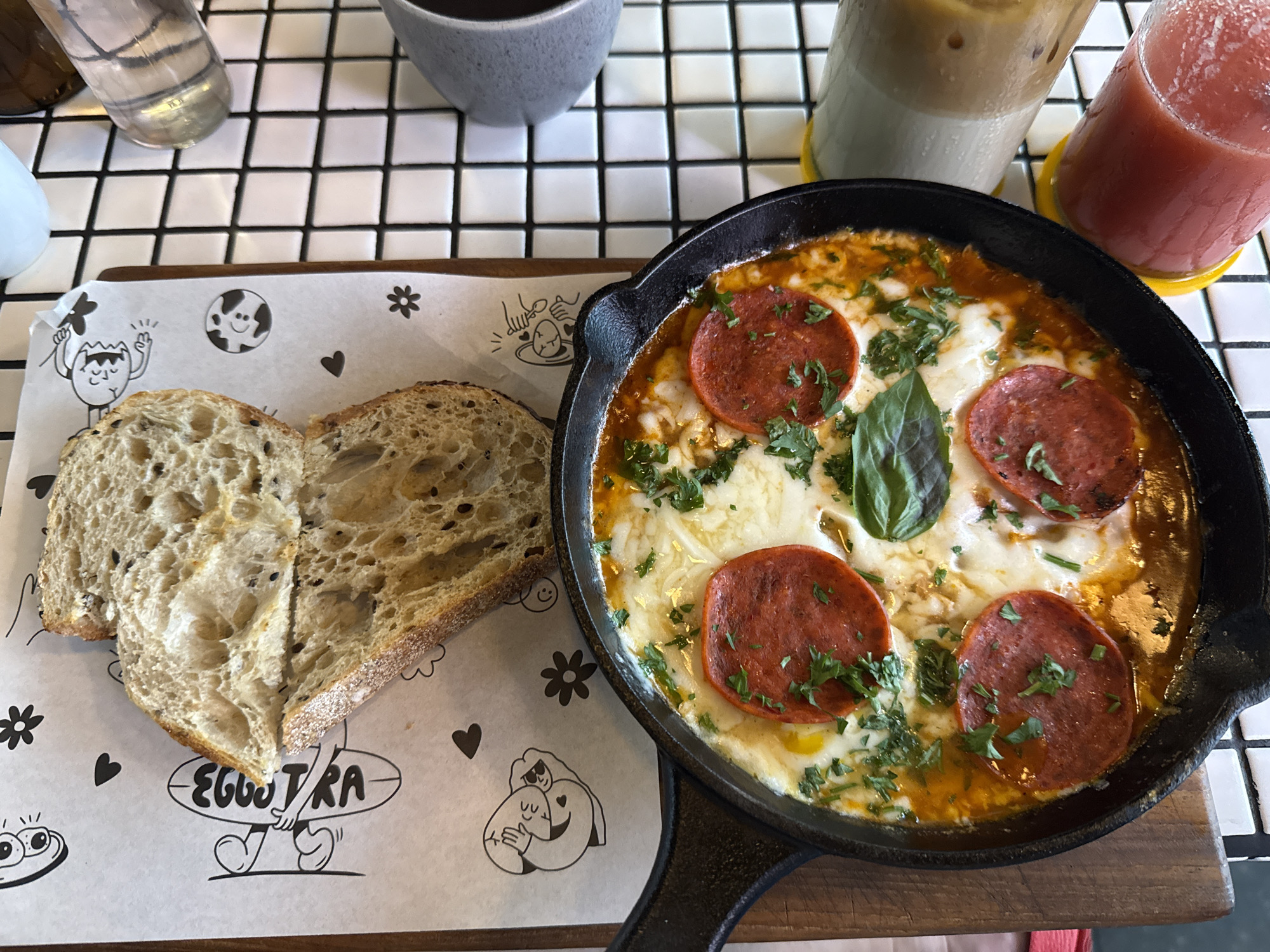 The image displays a delectable breakfast or brunch spread at a cafe or restaurant. The main subjects are a savory egg dish, artisanal bread, and several beverages, meticulously arranged on a wooden serving board over a black and white checkered tiled table.
At the center-right, a black cast-iron skillet holds a bubbling shakshuka-like dish. It features a bright red, tomato-based sauce generously topped with melted white cheese, four prominent slices of pepperoni, and a sprinkling of fresh green herbs, possibly parsley or cilantro. A single large basil leaf adorns the center, adding a fresh garnish. The dish appears hot and freshly prepared, with the cheese still gooey and the sauce simmering.
To the left of the skillet, two thick slices of light-colored, rustic bread, possibly sourdough or artisan whole-grain, are arranged to form a heart shape. The bread exhibits an open, airy crumb and has visible seeds or grains embedded within. These bread slices rest on a white paper liner, which is decorated with whimsical black line-art cartoons depicting various egg-themed characters, small hearts, and flowers. The brand name "EGGUTRA" is clearly visible in stylized text on the paper liner, held by cartoon hands.
In the background, several drinks are present. A tall glass contains a layered cold beverage, likely iced coffee or a similar milky drink, with ice cubes visible. Next to it, another tall glass holds a vibrant red or pink fruity drink, such as a juice or smoothie. Both tall glasses are placed on bright yellow coasters. Further back, a light gray ceramic cup and a clear glass of water are also visible.
The overall scene suggests a casual and inviting dining experience, likely during breakfast or brunch hours, with visually appealing food and drink presentations.