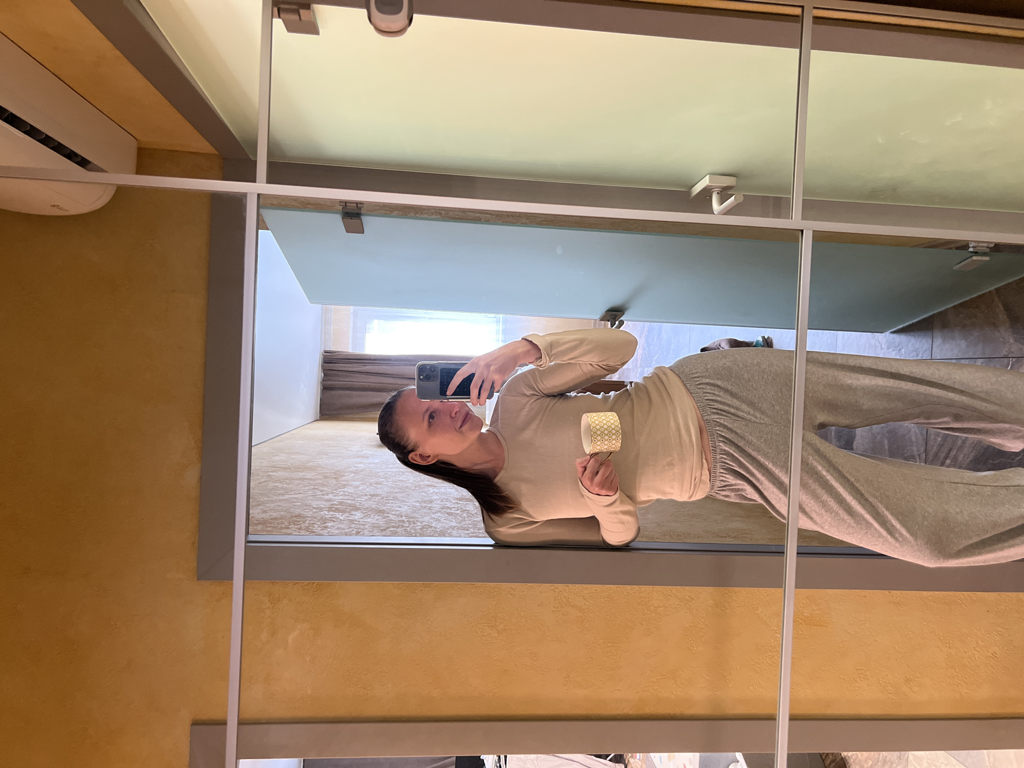 The image shows a person reflected in a multi-paneled mirror. The person is holding a smartphone in one hand, seemingly taking a selfie. They are wearing a light beige long-sleeved shirt and grey sweatpants. They are also holding a small paper cup.
The background reflected in the mirror shows a room with a window and curtain. The surrounding wall has a textured, golden-yellow paint. An air conditioning unit is mounted on the wall above the mirror.
There is no indication of the location being Khimki, Russia in the provided image.