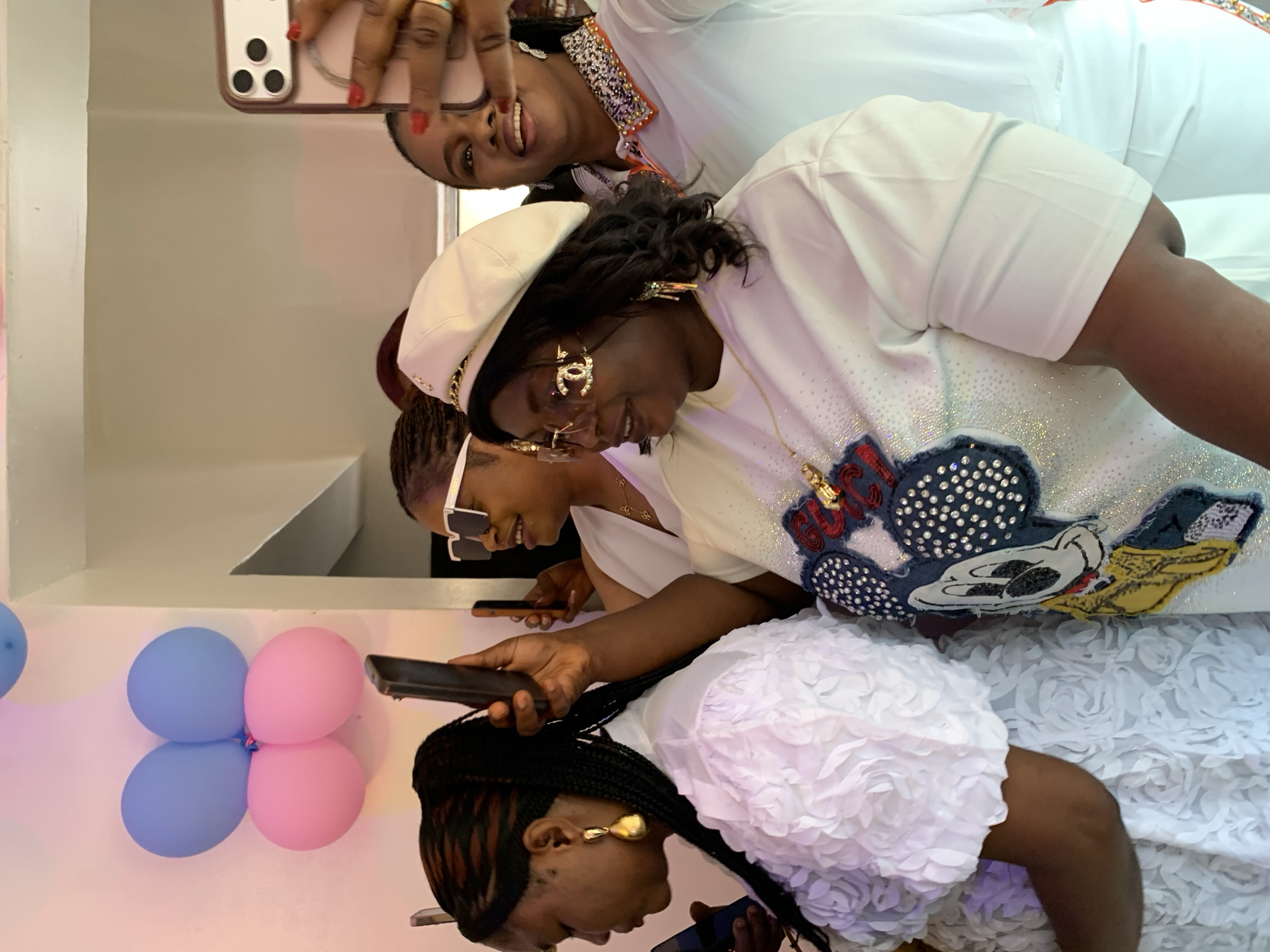 The image depicts a group of women at what appears to be a celebration, possibly a baby shower, given the presence of pink and blue balloons. The setting is indoors, with a neutral-colored wall and ceiling visible. The women are dressed in white attire, and some are holding up their phones to take pictures or record videos, suggesting a social event. One woman in the foreground is wearing a white beret and sunglasses with the Chanel logo. Another woman in the background is wearing white sunglasses. The overall atmosphere seems festive and joyful. The image does not provide any specific visual cues to pinpoint the location to the city of Jos, Nigeria.