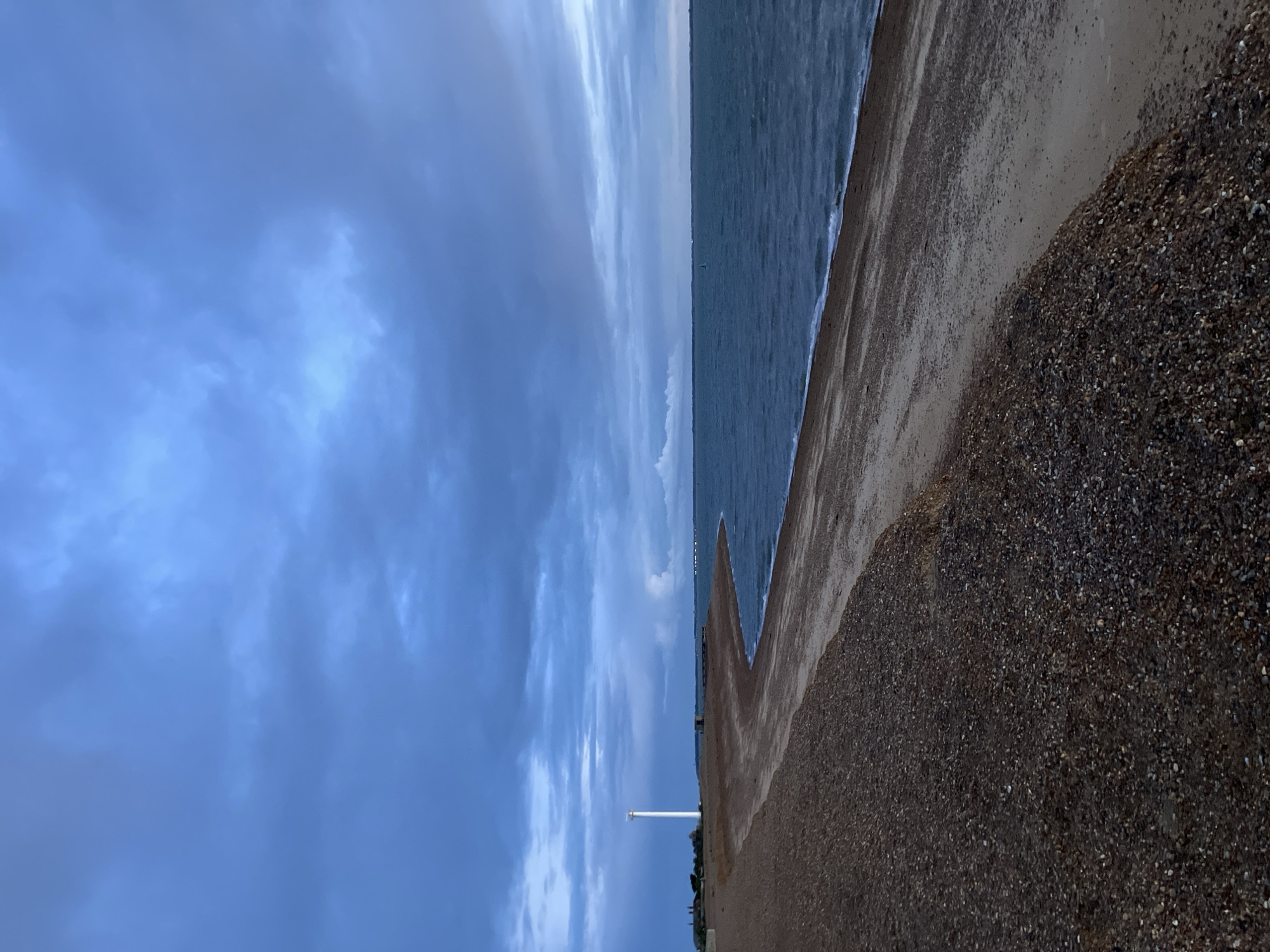 A serene coastal landscape is captured under a broad, overcast sky. The image portrays a tranquil scene featuring the sea meeting a pebble beach. The sky, occupying a significant portion of the upper frame, is filled with soft, textured clouds in various shades of blue and grey, indicating a cloudy or diffused light day.
A calm, dark blue-grey sea stretches horizontally across the midground, with gentle white-edged waves softly lapping at the shore. The foreground is dominated by a wide, textured pebble beach, transitioning from darker, coarser stones closer to the viewer to finer gravel and damp sand nearer the water's edge.
In the mid-distance on the beach, a tall, slender white pole or beacon stands vertically, a solitary man-made structure against the natural backdrop. Faint outlines of distant land or structures can be discerned along the far horizon line, where the sea meets the sky. No people or overt activities are visible, contributing to the peaceful and expansive atmosphere. The light is soft and even, consistent with an overcast day, without strong shadows to suggest a specific time of day. No visible text is present in the image. The setting evokes a sense of quiet solitude characteristic of a natural coastline, fitting the context of East Suffolk, United Kingdom.