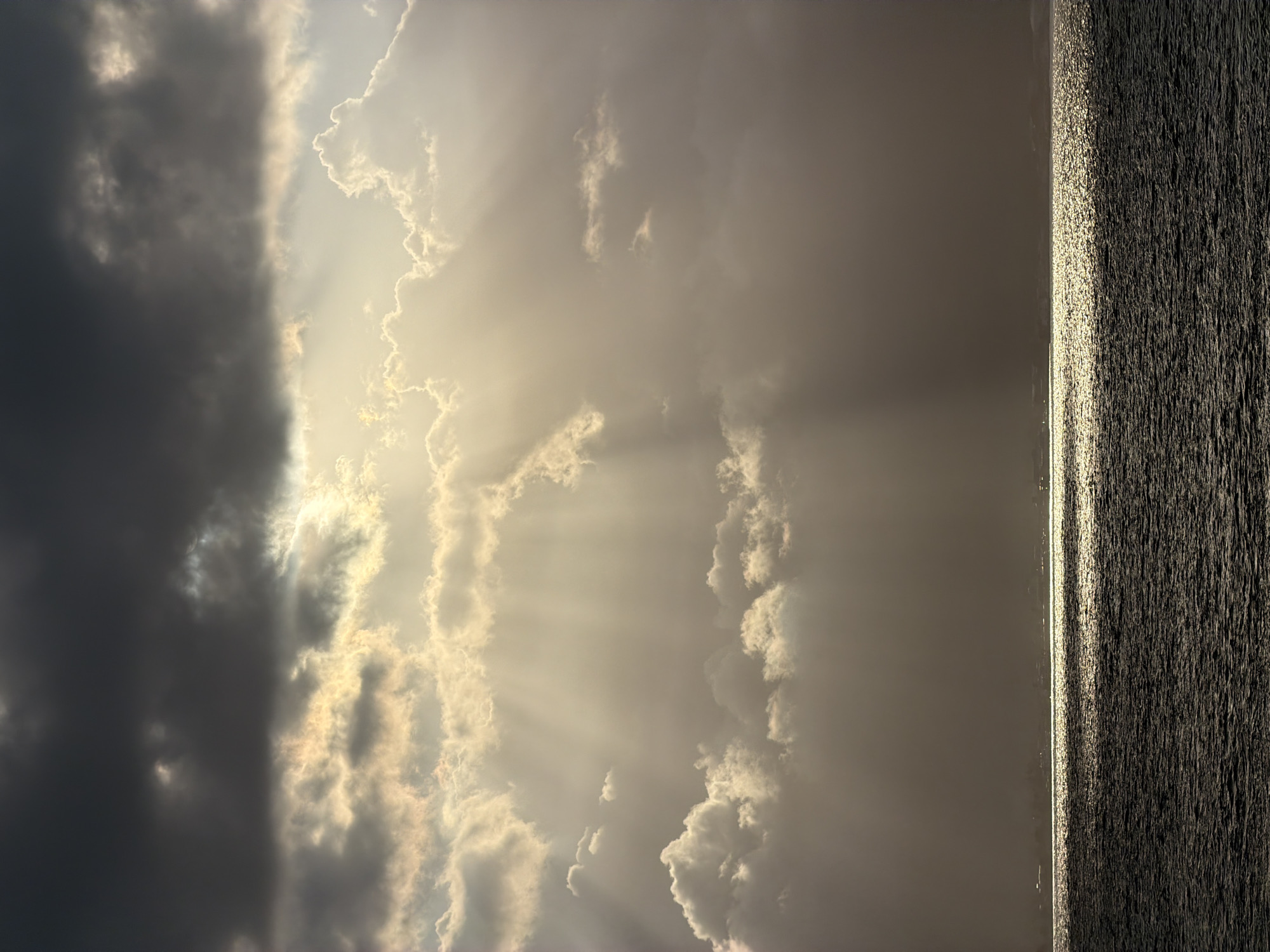 The image depicts a dramatic sky over a body of water. Dark, heavy clouds dominate the upper portion of the frame, with shafts of sunlight piercing through them, creating a crepuscular effect. These rays illuminate lighter, more scattered clouds below. The water in the foreground is dark and choppy, reflecting the light from the sky in a shimmering band along the horizon. A faint, hazy line of land with structures, possibly buildings, is visible in the distance along the horizon. The scene conveys a sense of natural power and atmospheric grandeur. The location is identified as Melaya, Indonesia, suggesting this is a coastal or offshore view near that area. No people or specific objects are discernible beyond the general elements of sky, water, and distant land.