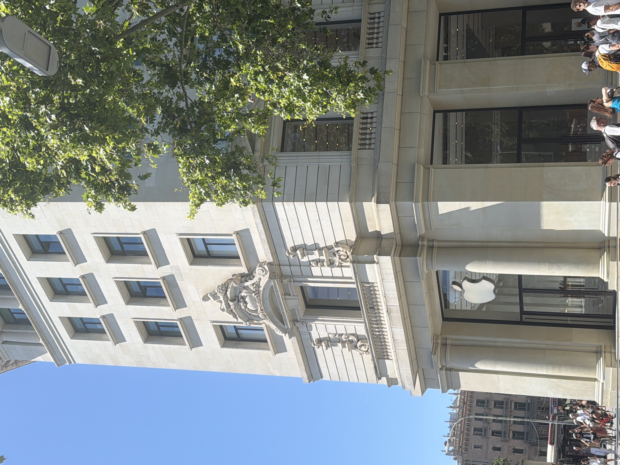 The image shows the exterior of the Apple Store located in Barcelona, Spain. The building is a large, ornate structure with many windows, and classical detailing. A large Apple logo is visible on the front of the store. A street light and a large tree overhang the building. There are people walking along the sidewalk and standing outside the store. Additional buildings are visible in the background. The sky is clear and blue.