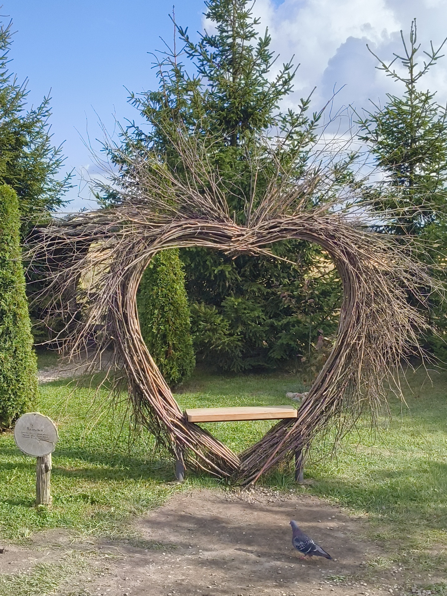 The image depicts an outdoor scene in what appears to be a park or garden. In the center of the frame, a large heart-shaped structure, meticulously crafted from intertwined branches, stands prominently. Within the heart, a wooden bench is positioned, suggesting a place for seating. The structure is surrounded by lush green grass and various types of trees, including coniferous varieties that frame the heart. To the left of the heart-shaped structure, a wooden post with a circular sign is visible, partially obscured by grass. In the foreground, a dirt path leads towards the heart, and a single pigeon is standing on the path. The sky above is blue with scattered clouds, and the lighting suggests it is daytime, likely with sunlight casting shadows. The location of Малахавецкі сельскі Савет, Belarus is referenced as context for this scene, implying this is likely a public space within that administrative region.