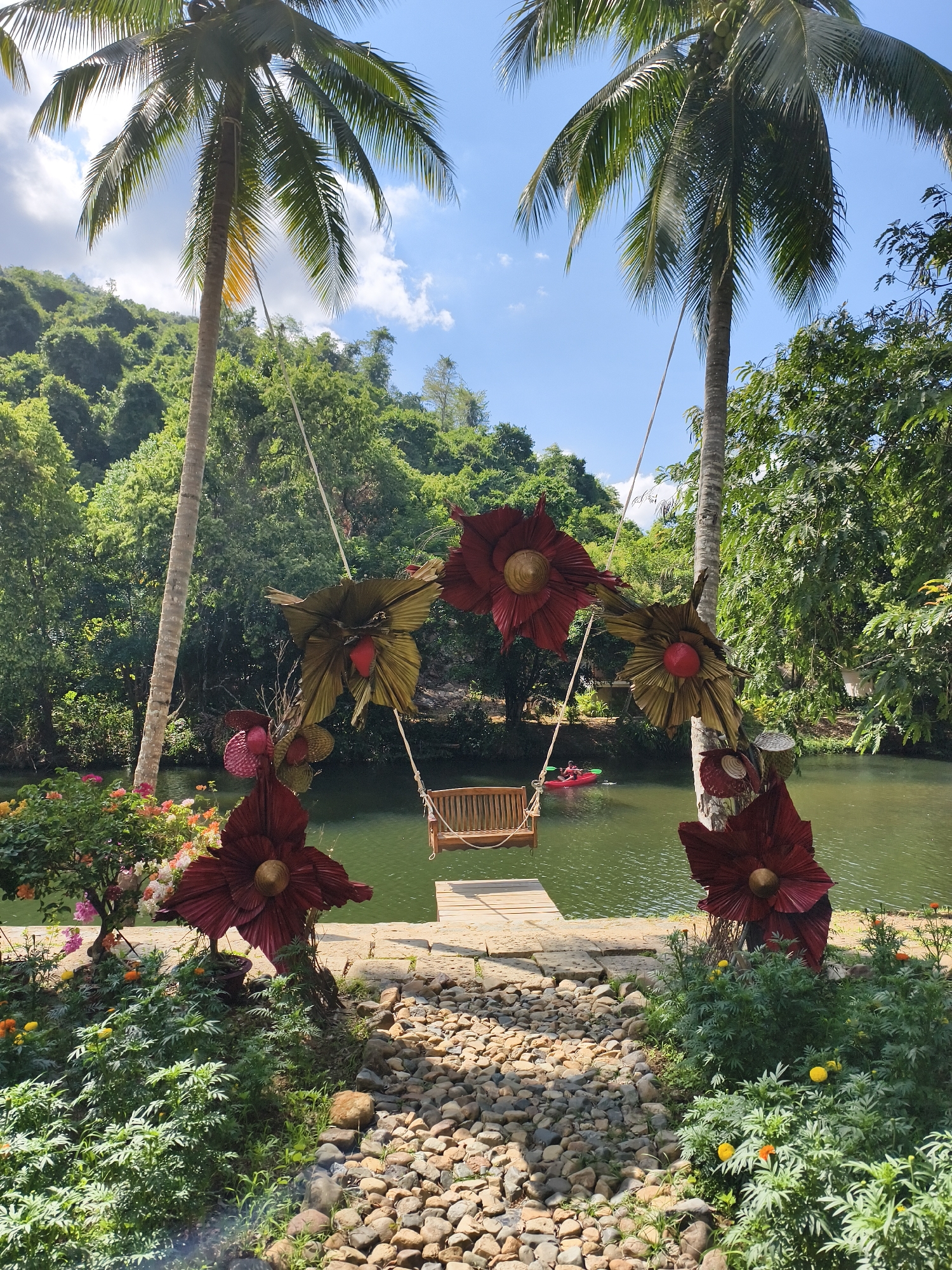 The image captures a scenic view in an unknown location. The foreground features a pebble-strewn path leading to a wooden swing hanging from the trunks of two palm trees. The swing is adorned with large, decorative floral arrangements in shades of red and gold. In the background, a serene river flows, with lush green vegetation covering the hillsides. A person in a red kayak is seen paddling on the river. The sky is partly cloudy, suggesting a bright, daytime setting.