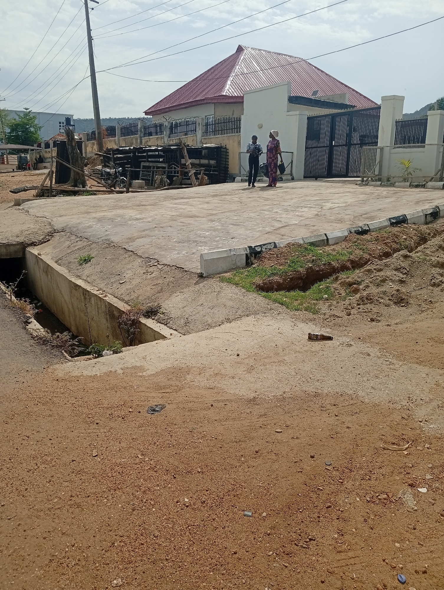The image captures a street scene in Adankolo, Nigeria. In the midground, two women are walking near a gated residence with a red roof. The property is secured by a metal fence and concrete pillars. To the left of the entrance, there is a pile of construction materials and debris. The foreground consists of a dirt road and a concrete drainage ditch. An overhead power line is visible against a cloudy sky.