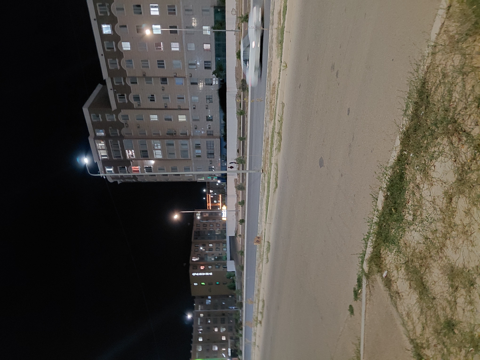 The image captures a nighttime street scene in Aktau, Kazakhstan, featuring several apartment buildings. The buildings are multi-story structures with numerous illuminated windows, suggesting residential occupancy. Streetlights are visible, casting light onto the road and surrounding area. A white car is in motion on the road, appearing as a blur due to its speed. Vegetation, including grass and small bushes, is present along the roadside and in front of the buildings. The overall environment appears to be an urban residential area at night.