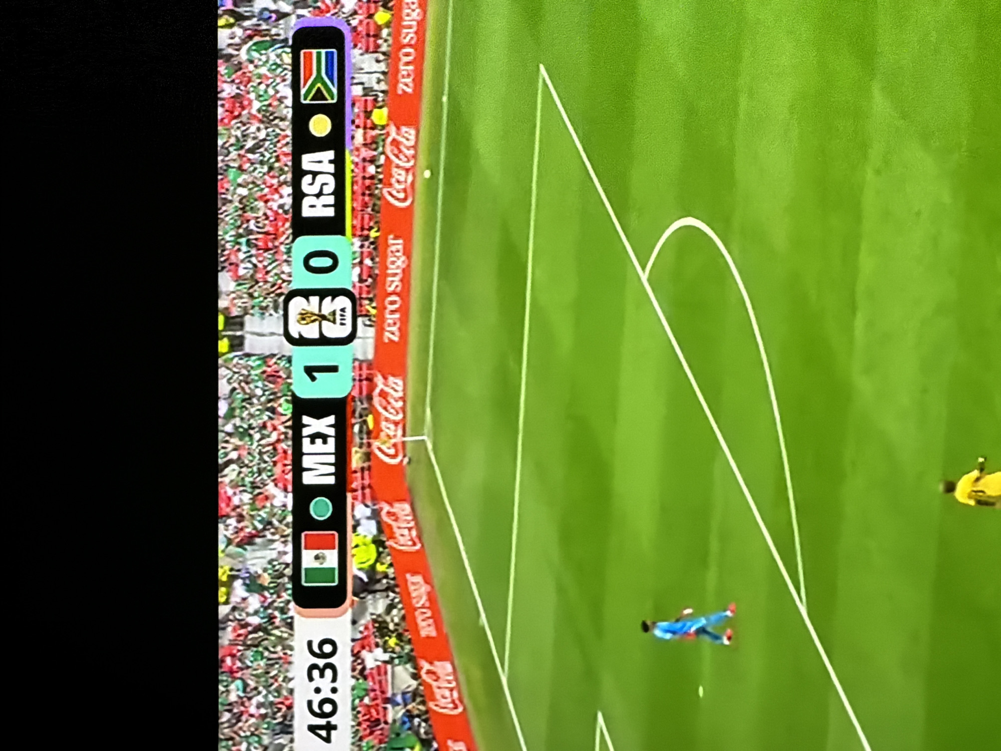 This is a still image of a soccer match displayed on a screen, likely a television or monitor, as indicated by the black border at the top and sides. The scene depicts a soccer field with white markings, including a penalty box. The scoreboard at the top of the image shows the time as "46:36". To the right of the time, it displays "MEX 1 0 RSA", with the Mexican flag next to "MEX" and the South African flag next to "RSA". A FIFA trophy logo is positioned between the scores. In the background, a crowd of spectators wearing red and green is visible. Along the edge of the field, there are prominent "Coca-Cola" advertisements, some with "zero sugar" text. On the field, two players are visible: one wearing a blue jersey and orange cleats, and another in a yellow jersey. The environment is a brightly lit soccer stadium.
