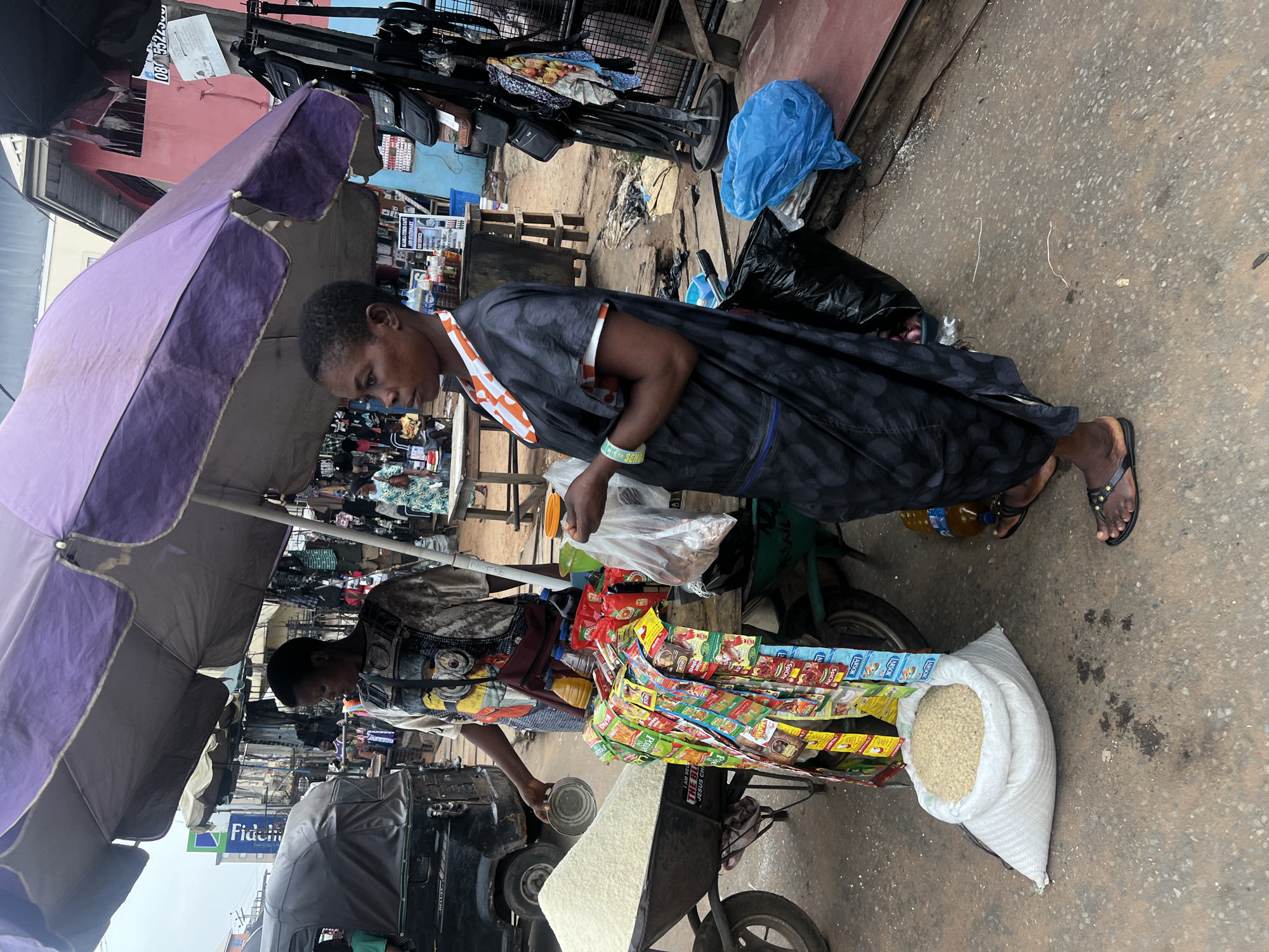 In Nkpor, Nigeria, a woman in a dark, patterned dress is buying a clear plastic bag of white rice from a vendor at a bustling street market. The vendor, standing under a large purple umbrella, measures rice with a metal cup from a large white sack on a three-wheeled bicycle cart. Numerous colorful packets of spices and seasonings decorate the cart. Nearby, another bag of rice sits on the ground. Various items, including clothing and bags, hang from overhead structures. A three-wheeled vehicle is parked close to the vendor. The street is littered with debris. Shop signs and other market structures are visible in the background.