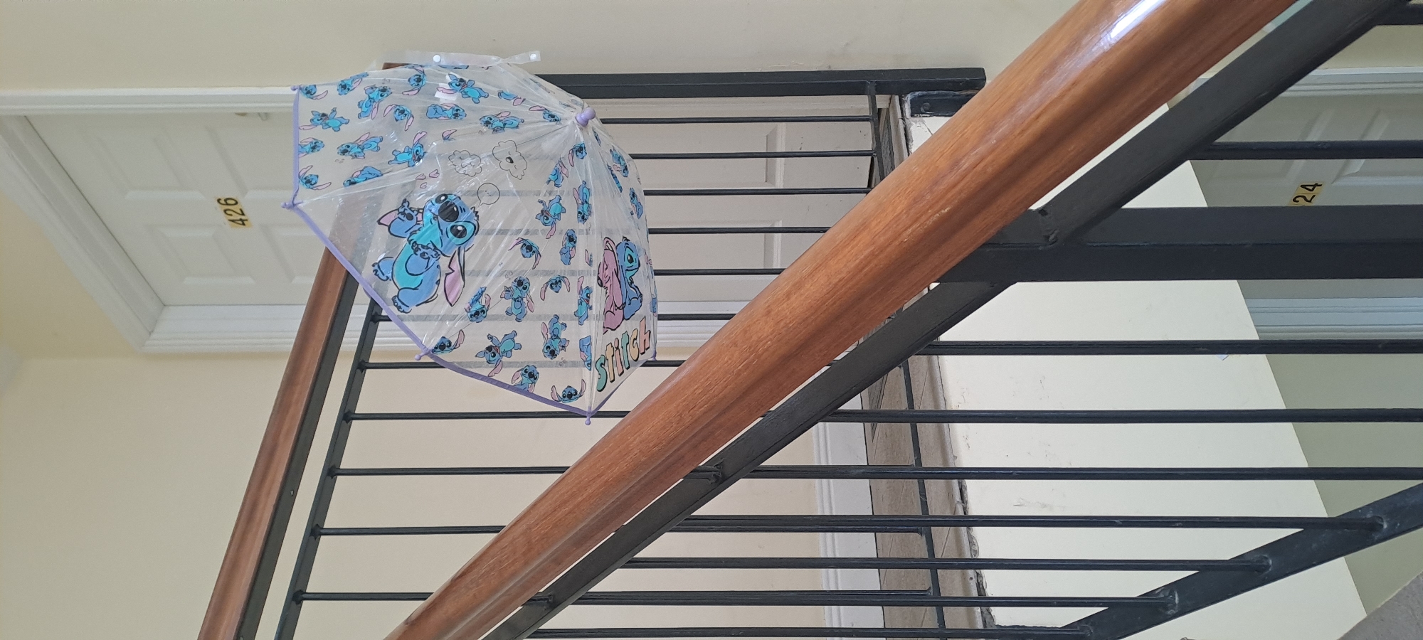 A transparent umbrella with a Stitch print is hanging from a black metal banister on a staircase. The umbrella is partially open and features multiple images of the character Stitch. In the background, a white door with the number "426" is visible. The staircase has a polished wooden handrail. A door with the number "24" is also visible in the background. The scene appears to be an indoor staircase within a residential building.