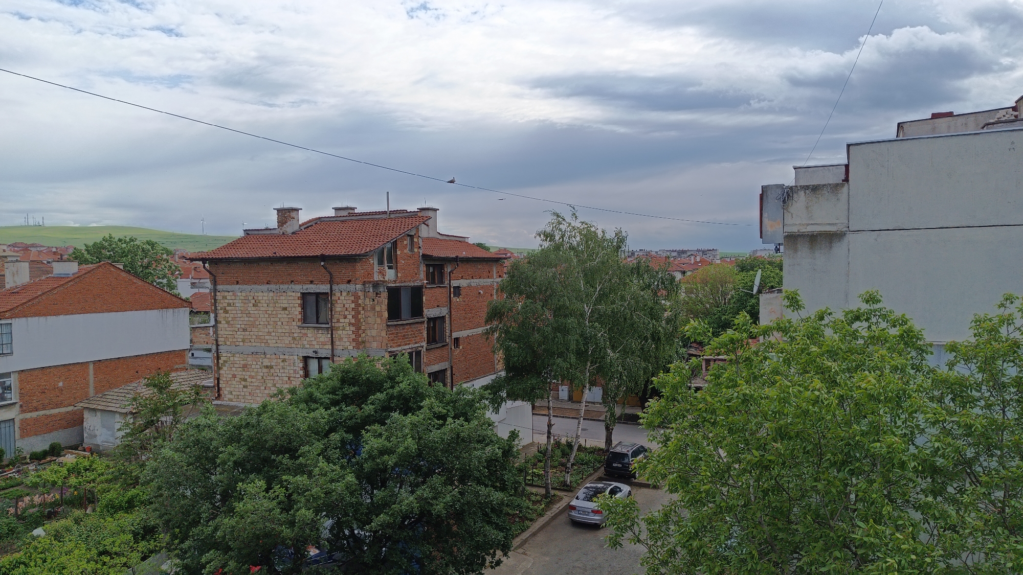 A medium-angle shot captures a view of an urban scene. In the foreground, lush green trees and foliage dominate the bottom portion of the frame.
Behind the greenery, a mixture of residential buildings is visible. The architectural styles vary, with some buildings constructed from red brick and others plastered in white. One partially finished structure has exposed brick and concrete blocks, revealing ongoing construction. A red-tiled roof is a common feature across the buildings.
Parked cars are visible along a narrow street nestled between the buildings and trees. Overhead, a utility line stretches across the frame. In the background, a green hill is visible.
The sky overhead is overcast with grey and white clouds.
The location appears to be Unknown, Unknown.