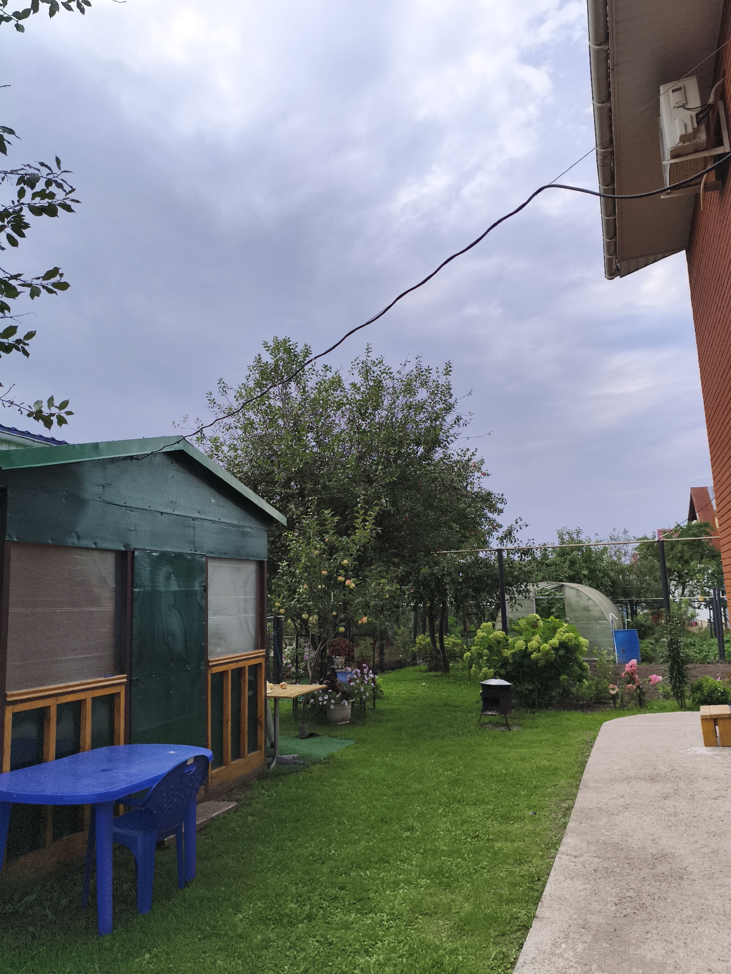 The image depicts a backyard scene in the city of Chaikovsky, Russia, on an overcast day. In the foreground to the left is a blue plastic table with a matching chair. Behind the table is a green wooden structure with a ribbed roof and translucent panels, appearing to be a shed or greenhouse extension. To the right of the structure, the scene opens to a lush green lawn. Further into the yard, a large green bush with bright green flowers is prominent, with a small metal barbecue grill to its left. Behind the bush, a transparent greenhouse structure is visible, and a blue barrel stands nearby. In the background, various trees and plants create a natural setting. On the far right of the image, the corner of a brick building is visible, with an air conditioning unit mounted on the exterior wall and electrical wiring running overhead. The sky is filled with heavy, gray clouds, suggesting a possibility of rain.
