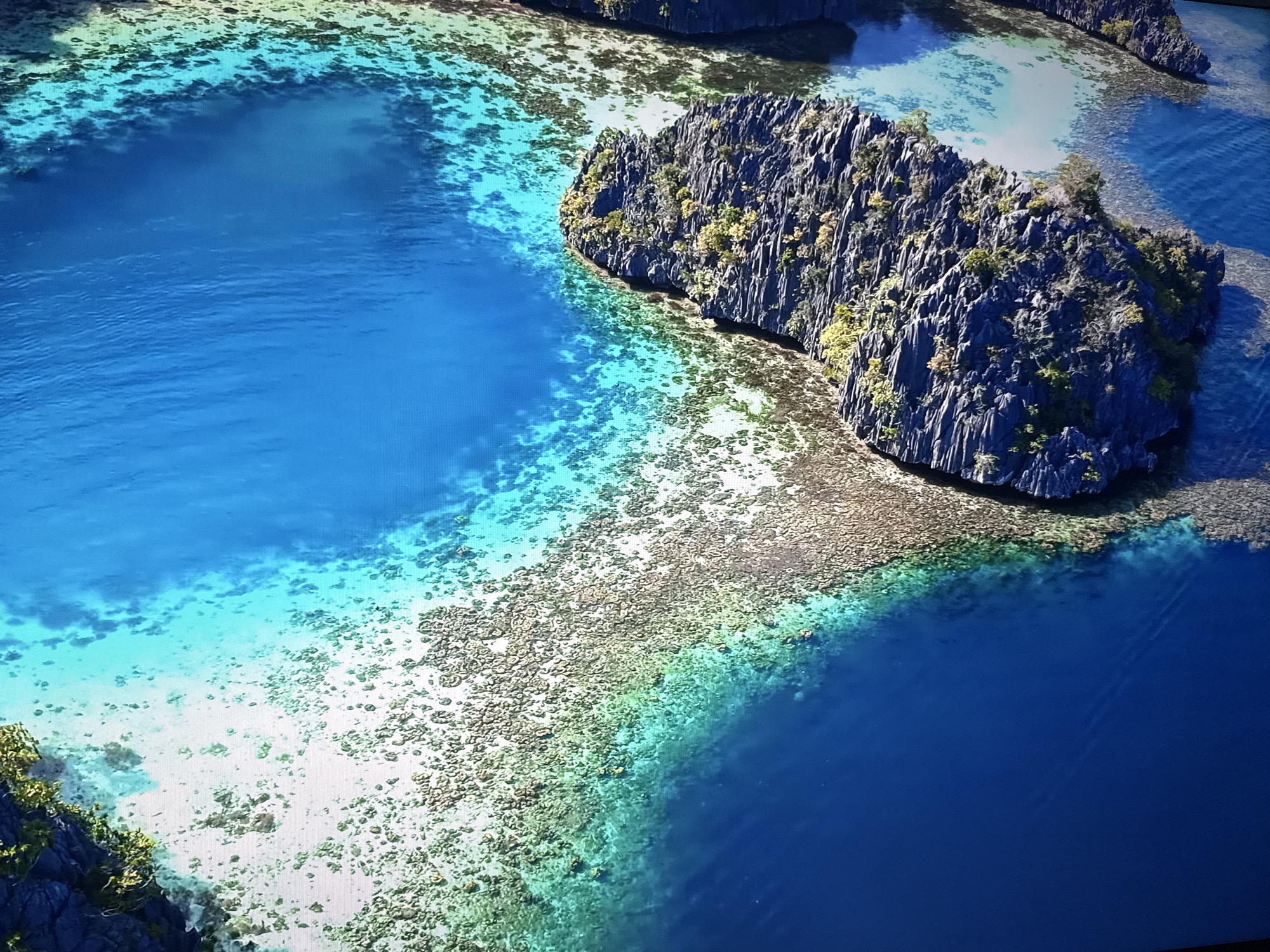 The image is an aerial view of a tropical island landscape. It features jagged, dark grey rocky outcrops covered in sparse green vegetation, jutting out into a vibrant turquoise and deep blue sea. The water near the shore is crystal clear, revealing a sandy bottom with scattered coral formations. In the distance, other similar islands can be seen. The scene is bathed in bright sunlight, creating a sense of a remote and pristine natural environment. There are no people or man-made structures visible in this image. The location is not in the city of Burgas, Bulgaria, as this depicts a tropical island.
