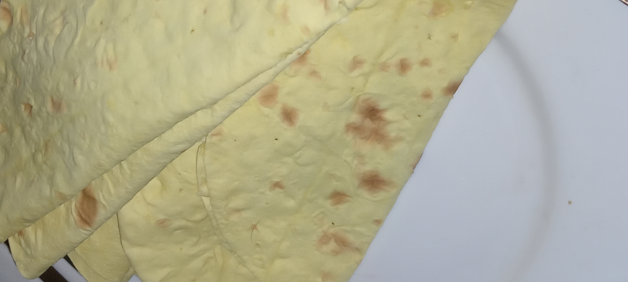 The image shows a close-up, overhead view of several folded flatbreads, likely lavash, stacked on a white plate. The flatbreads are pale yellow with irregular reddish-brown spots, indicating slight browning from baking. The texture appears soft and pliable. The plate is visible in the lower portion of the image and appears clean. There are no people or other objects in the frame. The scene is indoors, with neutral lighting that highlights the texture of the bread. There is no indication of the location being in Koviahy, Ukraine.