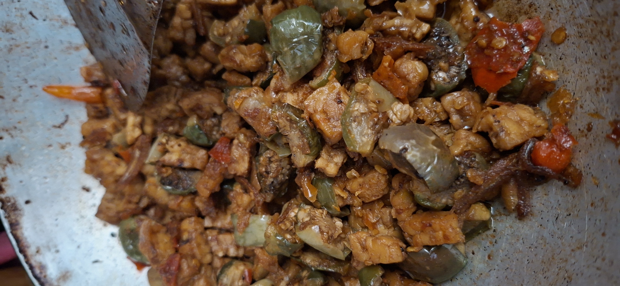 In a close-up shot, a dish of stir-fried tempeh and vegetables is being cooked in a metal wok. The tempeh is cut into small cubes and appears to be coated in a dark, glossy sauce. Mixed in with the tempeh are pieces of green bell pepper and what appear to be slices of eggplant. Small red chili peppers are also visible, adding a splash of color to the dish. A metal spatula is partially submerged in the food, suggesting it is actively being stirred. The wok itself shows signs of use, with a slightly worn surface. The background is out of focus, but the image suggests an indoor setting, possibly a kitchen or food stall. The dish is characteristic of Indonesian cuisine, and based on the presentation, it is likely being prepared in a street food stall or a modest eatery in Surabaya, Indonesia.