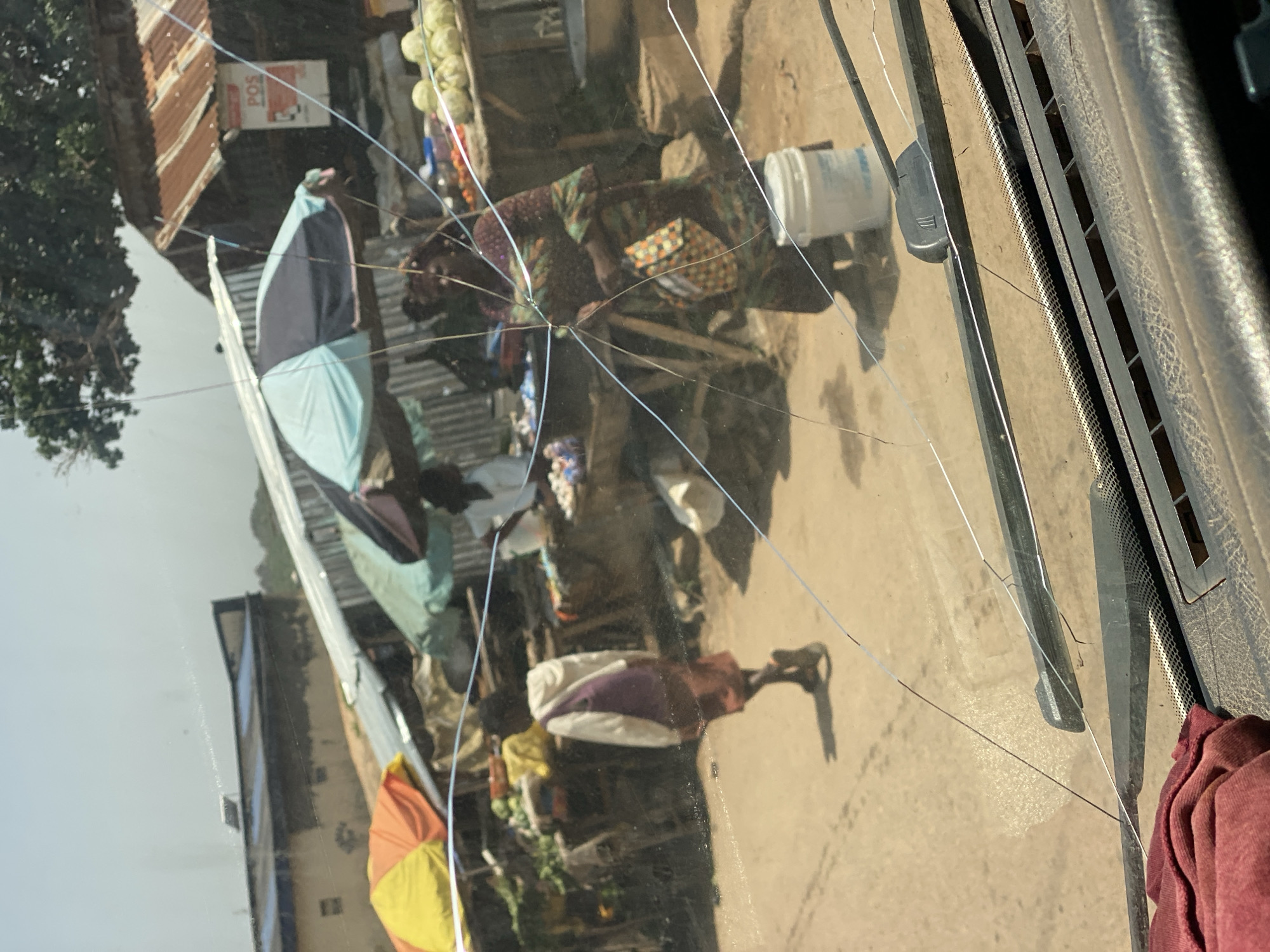 The image shows a street scene in Jos, Nigeria, as viewed through a cracked windshield. In the foreground, a woman in a colorful patterned dress is bent over, appearing to be sorting or arranging items. Beside her is a white bucket. Further into the scene, another person is partially visible, wearing a light blue and dark blue garment. A brightly colored umbrella, yellow and orange, is prominent in the lower left portion of the image.
The background reveals buildings with corrugated metal roofs and some structures that suggest market stalls or shops. A sign with the letters "POS" is visible on one of the buildings. The ground appears to be a dusty, unpaved surface, common in many parts of Nigeria. The lighting suggests it is daytime, with shadows cast on the ground, indicating the sun is out. The presence of market activities and everyday life implies this is a typical day in a bustling urban or semi-urban environment. The overall impression is one of economic activity and community interaction. The cracks on the windshield are a significant visual element, overlaying the scene and adding a layer of texture and disruption to the view.