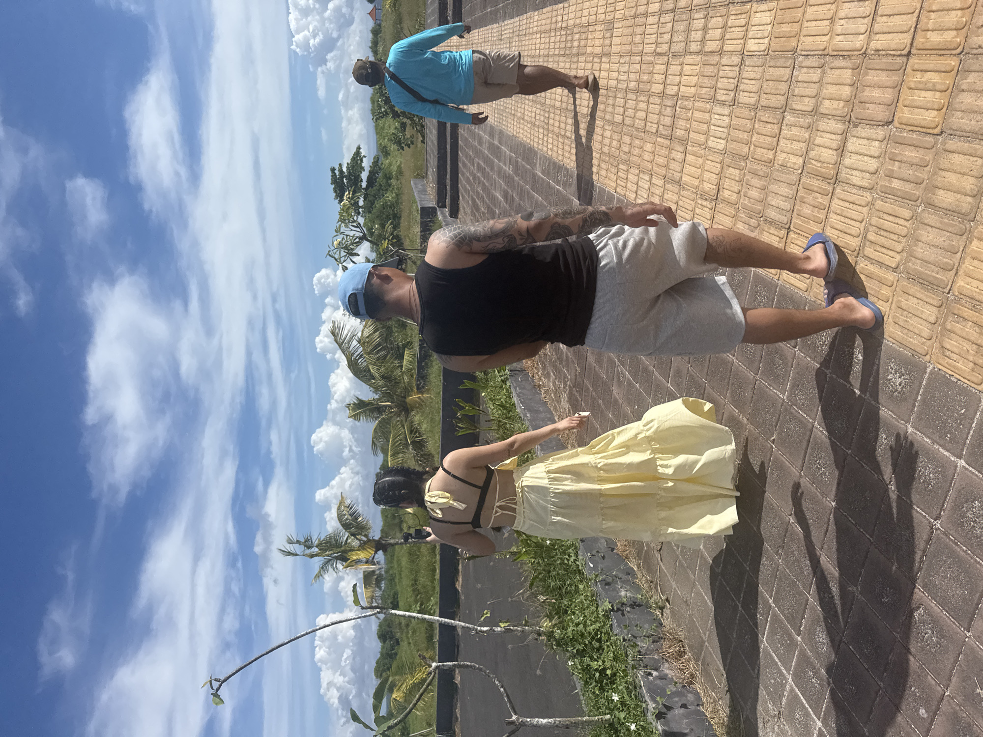 A group of people is walking on a brick pathway with a view of a tropical landscape and a partly cloudy sky in Nusa Penida, Indonesia. One person is wearing a yellow dress and has their back to the viewer, another is wearing a black tank top, gray shorts, and a blue baseball cap, and a third person is wearing a blue shirt and khaki shorts. The pathway is made of textured rectangular bricks, and there is green vegetation along the side. Palm trees and other tropical plants are visible in the background. The sky is blue with puffy white clouds.