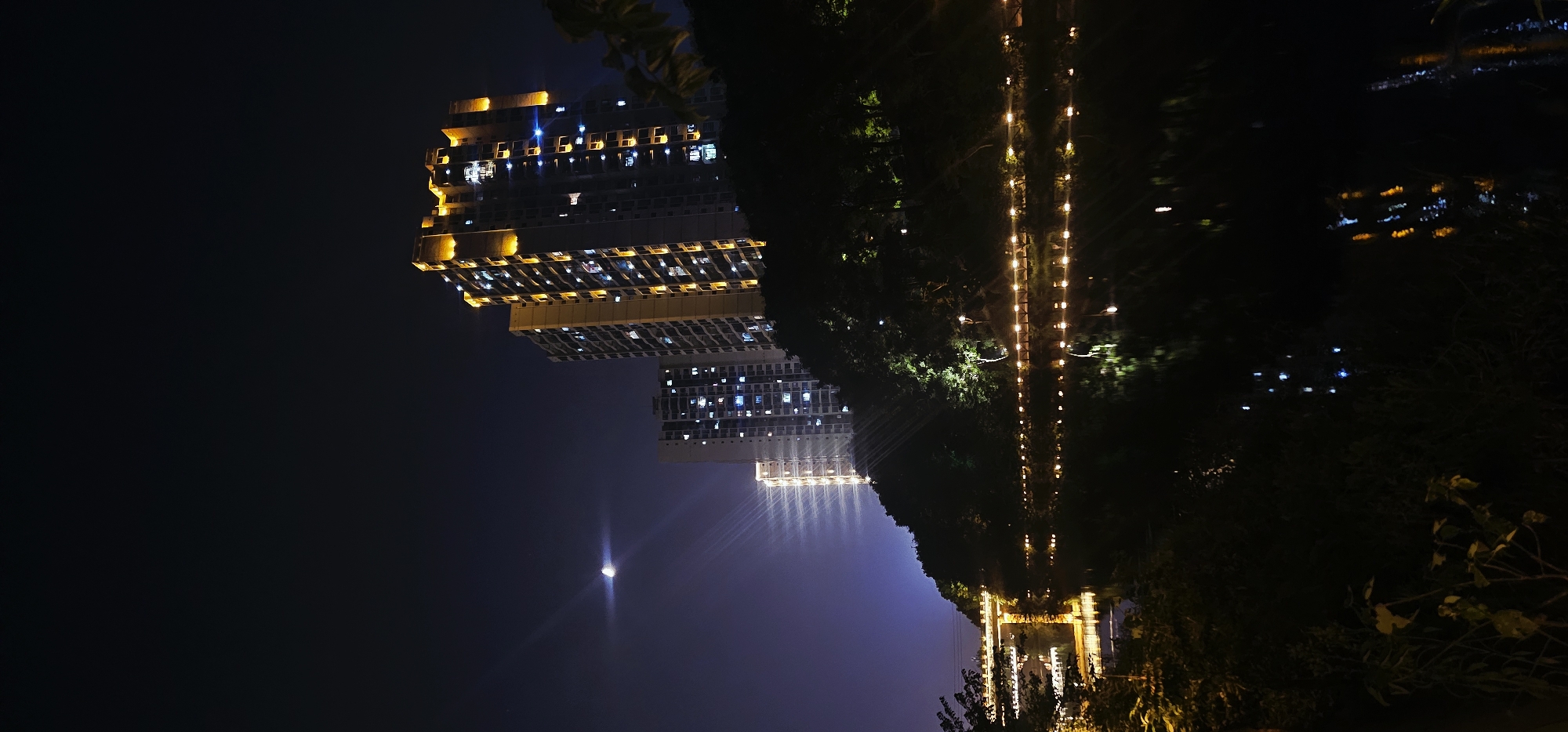This image depicts a nighttime scene in Bishan, China, featuring modern high-rise apartment buildings situated on a hillside overlooking a body of water. The buildings, illuminated with warm yellow and blue lights from their windows, dominate the upper portion of the frame. The tallest building, on the right, is prominently lit with accent lighting along its edges, highlighting its architectural features. A smaller, less illuminated building is visible to its left.
The hillside itself is densely covered with dark trees, with some strategically placed lights creating an ambiance along a pathway or promenade that runs along the water's edge. These lights, reflecting on the dark, still water, create a shimmering effect. A bridge, also illuminated with lights, spans across the water in the middle ground on the left side of the image.
In the sky, the moon is visible as a bright crescent, accompanied by distinct light rays or atmospheric effects emanating from it and also from the lights of the buildings. The sky transitions from a deep, dark blue at the top to a lighter, purplish hue closer to the horizon, possibly due to light pollution. The foreground consists of dark foliage, adding depth to the scene and framing the view of the city lights and water. No people are discernible in the image.
