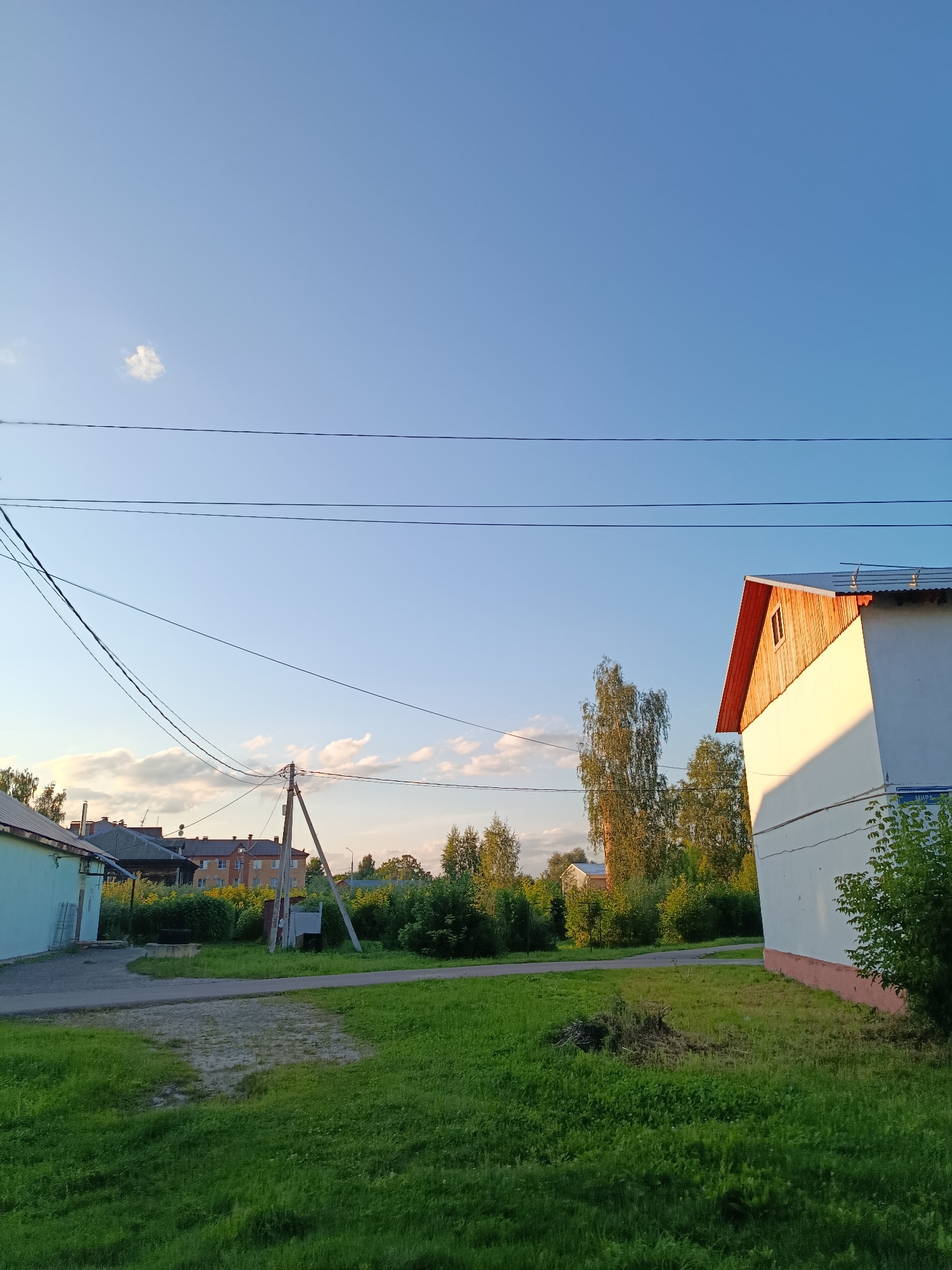 The image depicts a clear sky with a few wispy clouds. In the foreground, a patch of green grass with some dry brush is visible. To the right, a white building with a red-trimmed gable roof, featuring a small window, is illuminated by the setting sun. A street sign, partially visible, reads "МИРА", indicating Mira Street. In the middle ground, a paved road curves past a light blue building on the left. Further back, a collection of trees and foliage separates the foreground from a line of residential buildings. A wooden utility pole with visible wires stands near the road. The scene appears to be a residential area in the city of Vereya, Russia, during the late afternoon or early evening. There are no people or animals present in the image.