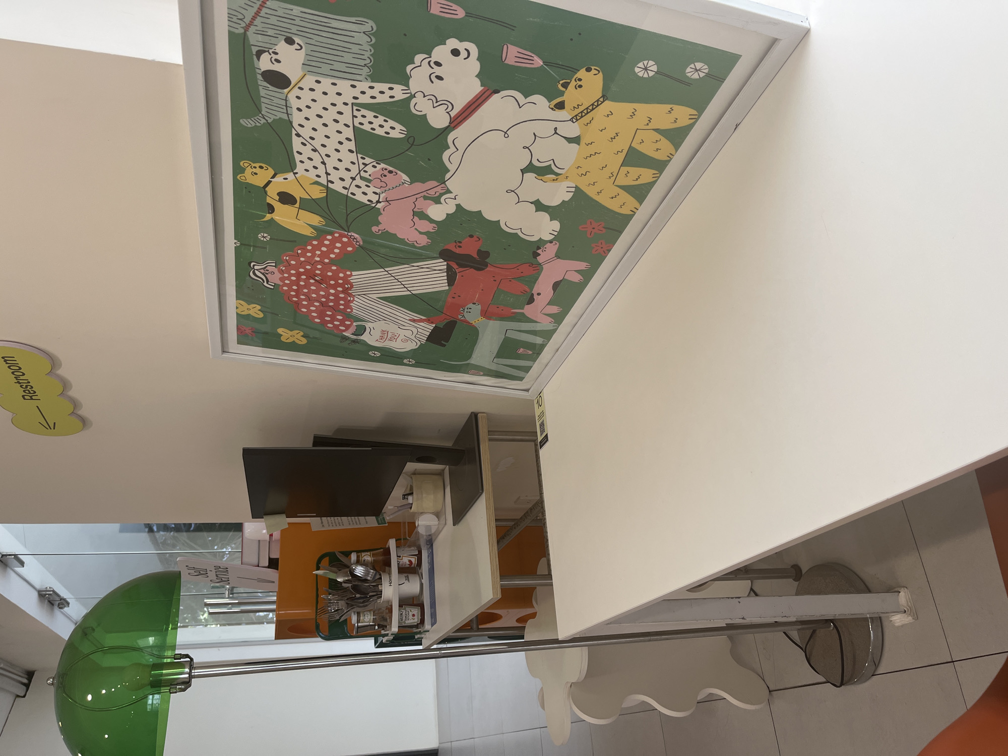 The image shows a whimsical framed artwork of dogs and people on a green background, hanging on a white wall. Below, a service area with a green lamp, orange utensil container holding silverware and condiments, a white shelf with a screen or tablet on it, and a white table with decorative, scalloped edges are visible. A restroom sign with a yellow cloud and arrow is mounted on the wall near the artwork. The scene appears to be in a commercial setting.