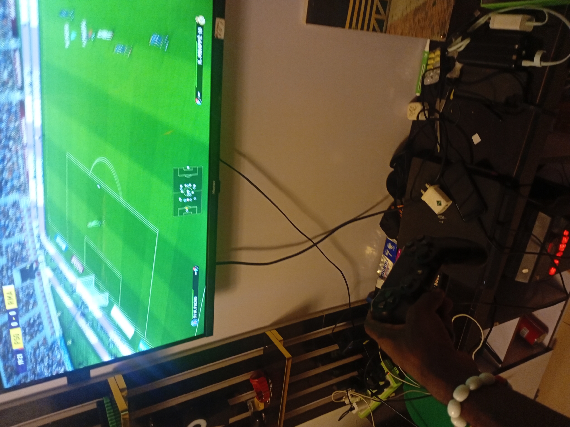 An indoor scene depicts a person engaged in playing a football (soccer) video game on a large television screen. The screen dominates the left side of the frame, displaying a vibrant green virtual pitch with players, goalposts, and stadium elements. The game's user interface is visible, showing "PSG 0 - 0 RMA" as the score, "19:21" as the game time, and player names such as "K. MBAPPE 80" and "V. JR. P/PHO." A mini-map of the pitch is also displayed in the lower right corner of the screen.
In the foreground, a person's arm, adorned with a white beaded bracelet, holds a black game controller, likely a PlayStation controller, indicating active gameplay. To the right of the television, a dark-colored table or desk is cluttered with various electronic devices, chargers, adapters, and tangled cables. Notable items on the table include a white power adapter with a possible "Mi" or "Xiaomi" logo and a partially visible "acacare" branded water bottle. The wall behind the setup is a light, plain color, with a black and yellow striped decoration visible in the upper right background. The overall setting suggests a dedicated gaming area, possibly in the evening or night due to the artificial lighting.