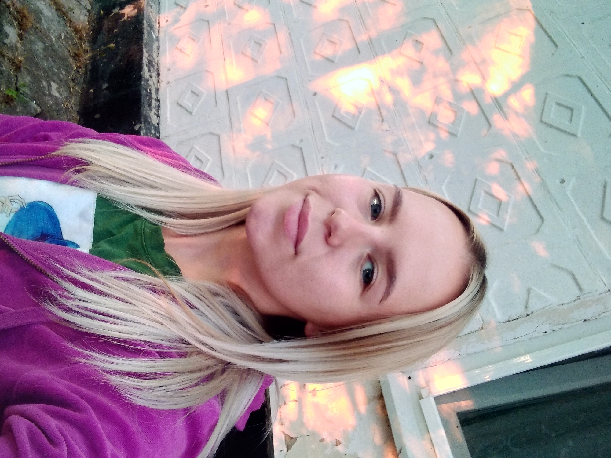 In Radisne, Ukraine, a young woman with blonde hair and blue eyes poses for a selfie in front of a textured, light-colored wall. The wall features a repeating pattern of diamond shapes, some of which are illuminated by bright, warm light, suggesting the setting sun. The woman is wearing a green t-shirt with a graphic print and a purple hooded jacket. The background also includes a window frame and a dark, rough surface at the bottom, possibly a stone or concrete ledge. The overall impression is of an outdoor portrait taken during golden hour.