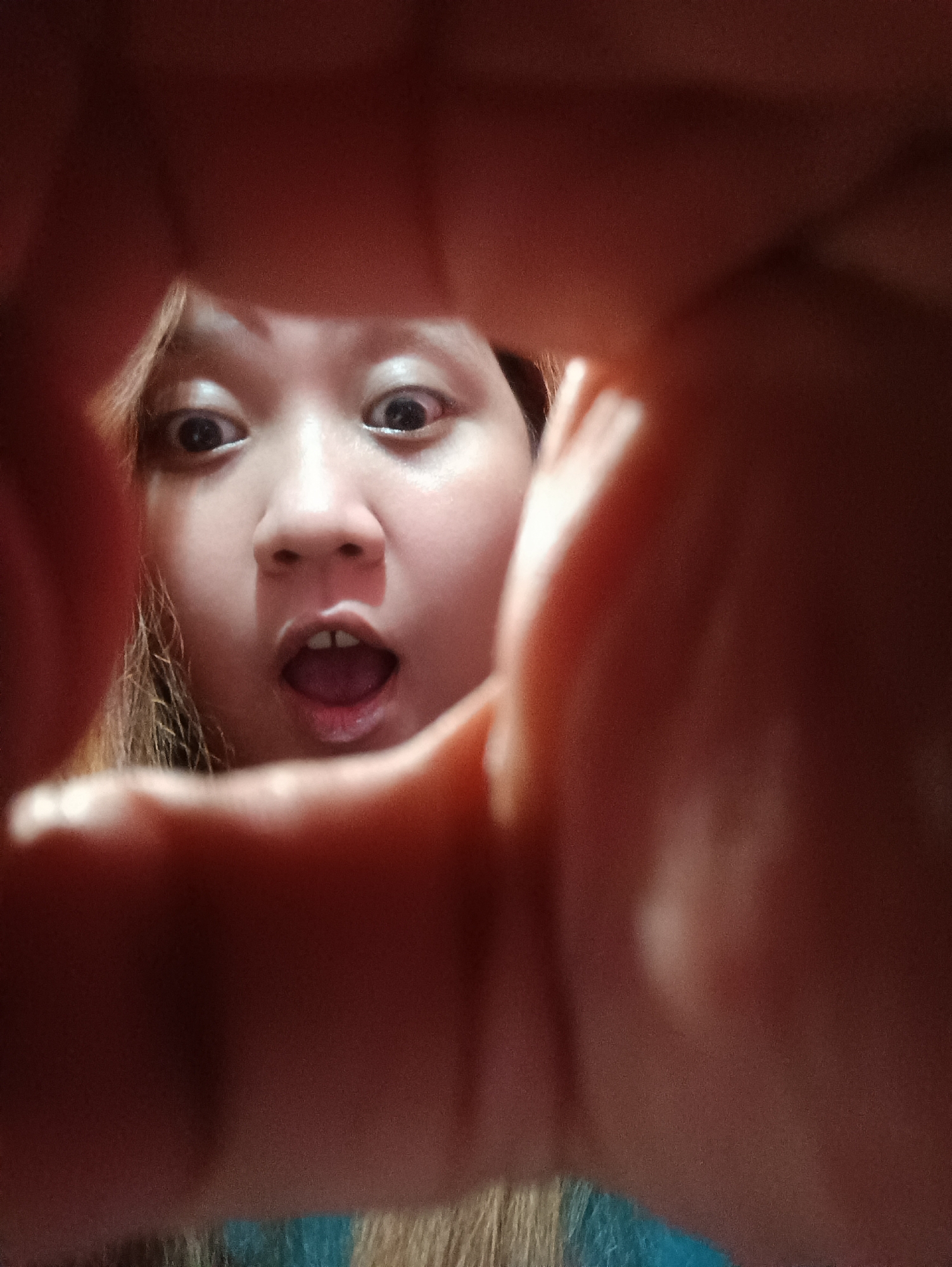 A close-up, selfie-style shot captures a young person's face peeking through fingers that frame the central subject. The person's eyes are wide open, and their mouth is agape, suggesting surprise or excitement. The background is dark and out of focus, with hints of blonde hair and a blue garment visible at the bottom of the frame. The lighting is dim and appears to come from the front, illuminating the person's face. The image was likely taken indoors in a residential setting in Depok, Indonesia.