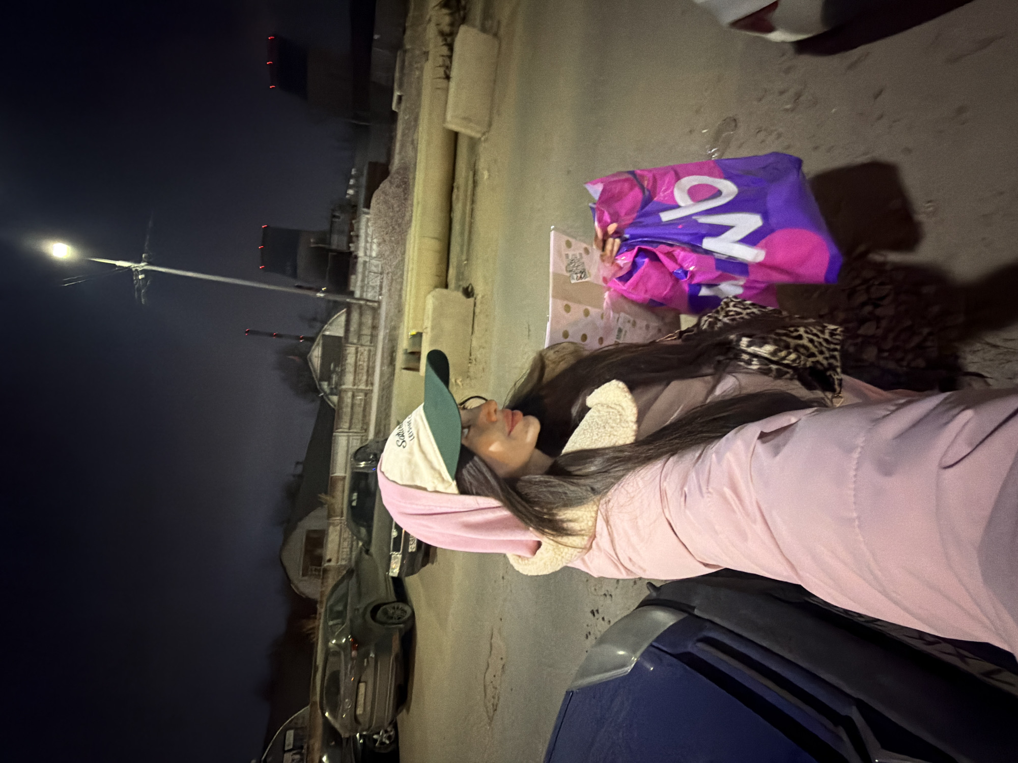 The image depicts a nighttime urban scene, likely a parking or industrial area, in Saint Petersburg, Russia. A female subject is visible in the foreground, positioned slightly right of center, looking towards her right in profile. She has long, dark hair and wears a light pink padded jacket with a cream-colored fleece lining, a pink hoodie, and a cream-colored baseball cap with a dark green brim that reads "SATURDAY LEISURE CLUB". In her right hand, she holds a purple plastic shopping bag with "доб" (DOB) written in white capital letters, indicative of the Russian supermarket chain "Доброцен". Her left hand holds a small, folded, polka-dotted item. The immediate foreground on the left shows the partial front section of a dark blue vehicle.
Behind the subject, a dark grey SUV (possibly a Mazda CX-5) and a dark sedan are parked on a paved surface. Further back, a pile of gravel is visible alongside several large concrete barrier blocks. The background features a tall street lamp with a bright luminaire, providing the primary light source for the scene. Beyond this, indistinct buildings, including industrial or utilitarian structures, are present. To the far right, large, dark structures with multiple red lights on their upper sections are visible, consistent with industrial facilities. A faint red-and-white striped chimney stack is discernible in the distant left background. The sky is dark, indicating night.