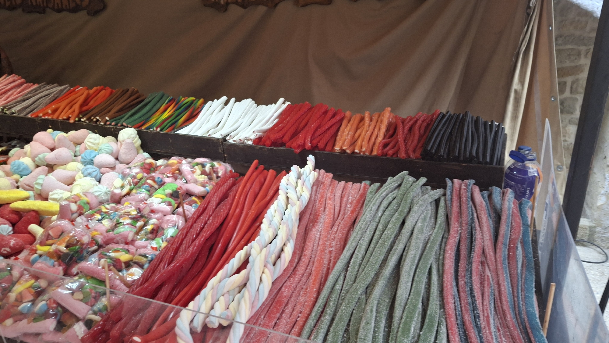 The image shows a vibrant display of assorted candies at a market stall, likely in the city of La Fresneda / la Freixneda, Spain, given the stone wall in the background which is characteristic of the region. The foreground is dominated by rows of colorful, twisted licorice-like candies. These candies are arranged in distinct lines, featuring various colors such as red, pink, green, blue, and black. Some of the candies appear dusted with sugar. To the left and slightly behind the licorice, there are piles of smaller, pastel-colored candies, some of which resemble meringue kisses and others are packaged in clear plastic bags, revealing a mix of smaller, brightly colored candies. In the background, a tarp-like fabric, possibly part of the stall's awning, hangs loosely. A blue plastic bottle with a label is visible on the right side of the display. The overall impression is that of a sweet shop or a market vendor specializing in confectionery.