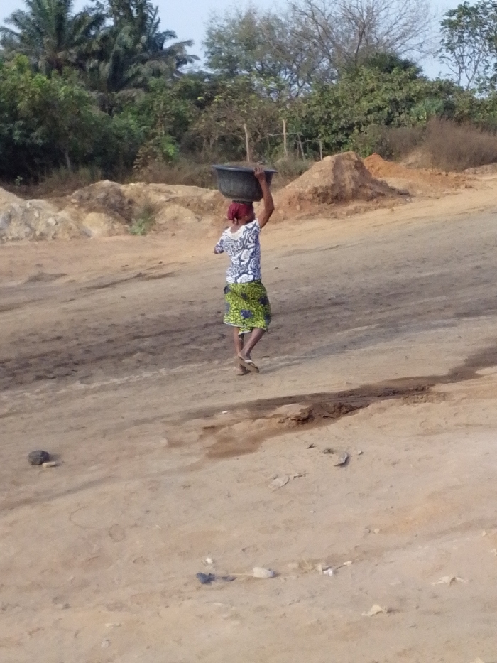 A lone individual is depicted walking on a wide, unpaved dirt road in Hoche, Nigeria. The subject, an adult female, is viewed from behind, positioned in the lower-left to mid-right quadrant of the frame, walking towards the right side of the image. She wears a reddish head covering, a white top with a dark patterned design, and a green, yellow, and blue patterned wrapper skirt. A dark-colored basin or bowl is balanced on her head, supported by her raised hands. She is wearing light-colored flip-flops. The ground surface consists of compacted light brown earth with visible tire tracks or footpaths, and several small rocks and debris are scattered across it. A shallow, dark-colored depression or rut spans horizontally across the mid-ground. In the background, multiple irregular mounds of light brown earth or sand are visible. The far background features dense tropical vegetation, including tall palm-like trees on the left and various deciduous and evergreen trees and bushes under a light, hazy sky.