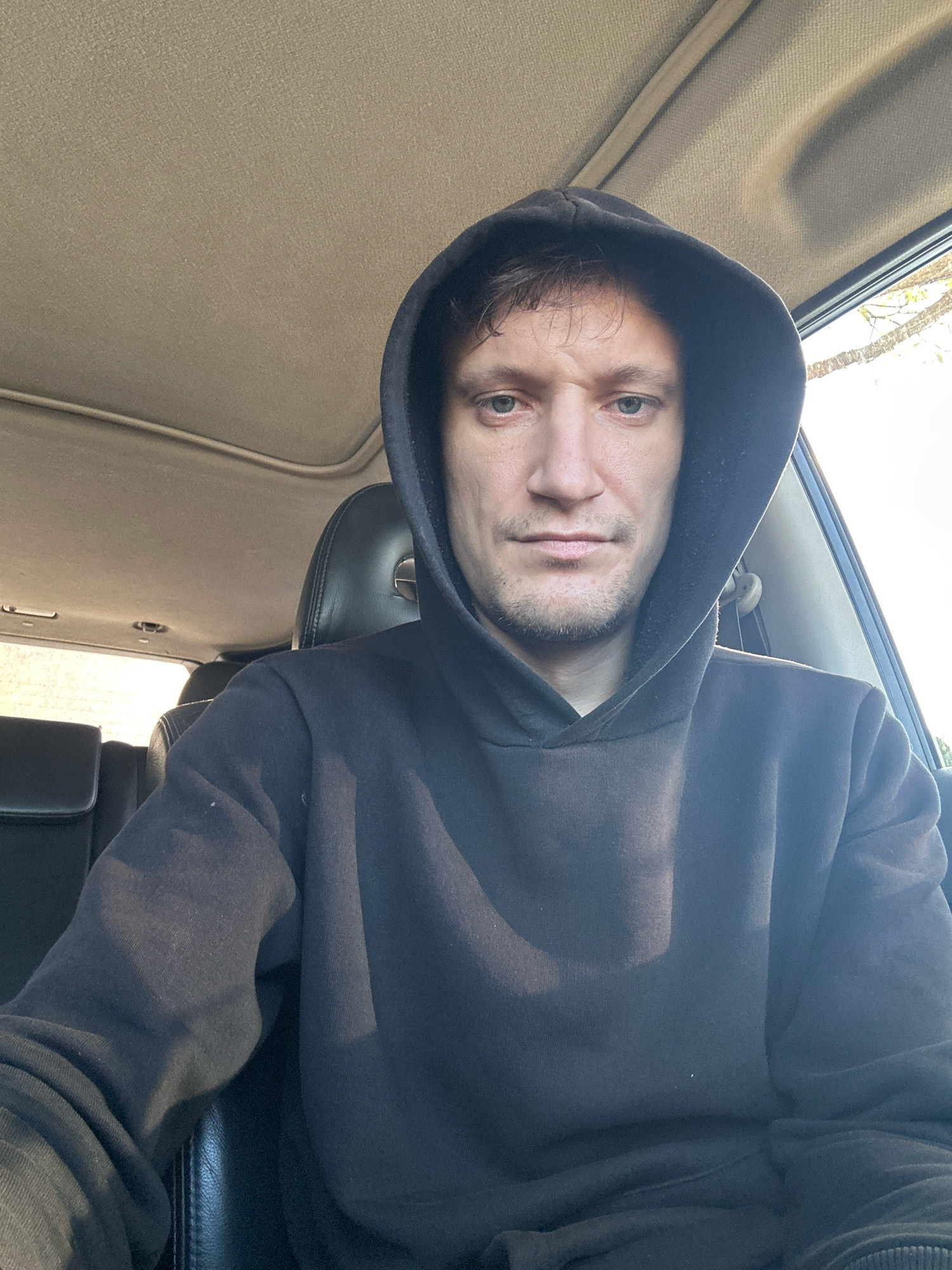 A man wearing a black hooded sweatshirt is seated in the driver's seat of a vehicle. The interior of the car is visible, including the headliner and seats. The man is looking directly at the camera. The sun is shining, casting light into the vehicle. The background outside the car appears to be trees and foliage. The image was taken in Makiv, Ukraine.
