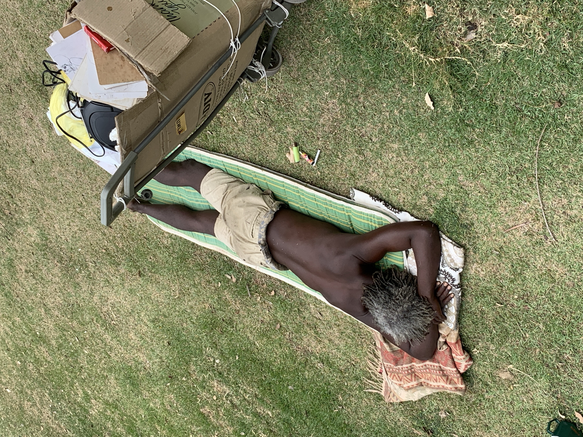 Overhead shot of an African-American male lying prone on a green striped mat on grass, with his head on a patterned cloth. He is wearing tan shorts and has grey dreadlocks. A utility cart filled with boxes and items such as a black bag and papers sits near his feet. An electronic cigarette and cartridges are placed beside him on the grass. This image was taken in Tel-Aviv, Israel.