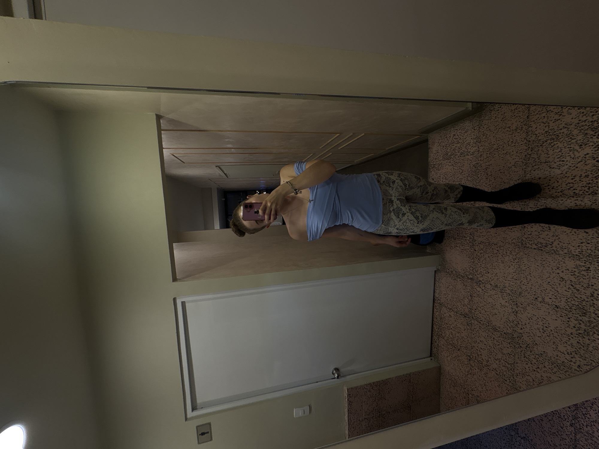 A woman in a blue tank top and patterned leggings takes a mirror selfie in a room with light beige walls, a white door, and a speckled floor. The location is likely in Mexico City, Mexico, based on the potential cultural elements within the room’s architecture. The woman’s face is obscured by her phone. She is wearing jewelry on her left wrist.