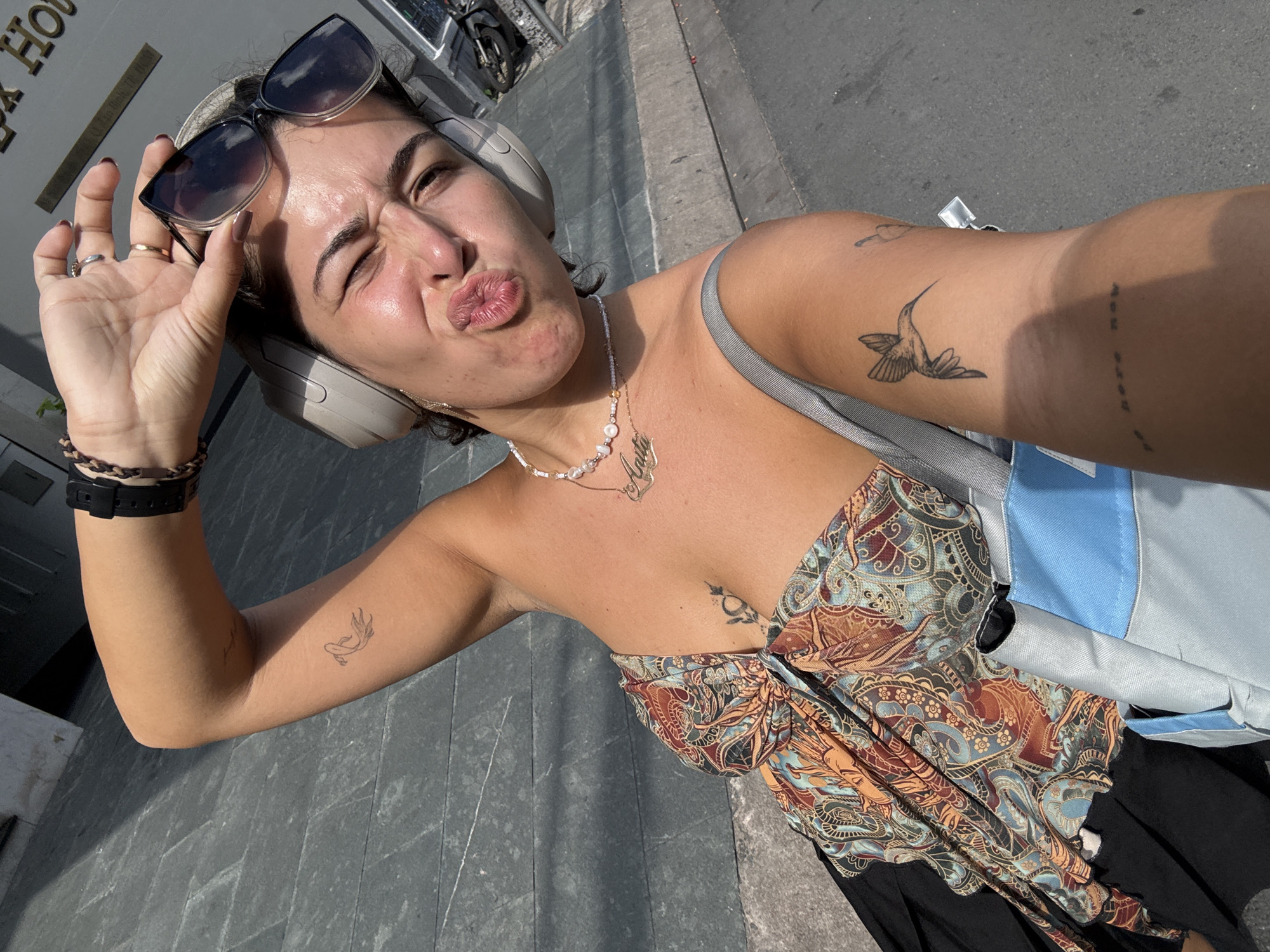 The image shows a young woman with headphones on, taking a selfie. She is wearing a colorful patterned top and black skirt. She has several tattoos on her arms and chest. She is holding sunglasses with her right hand and making a duck face. The background appears to be a city street with a building and a motorcycle visible. The ground is paved with gray stone tiles. The lighting suggests it is a sunny day. The location is identified as Thuận An City, Vietnam.