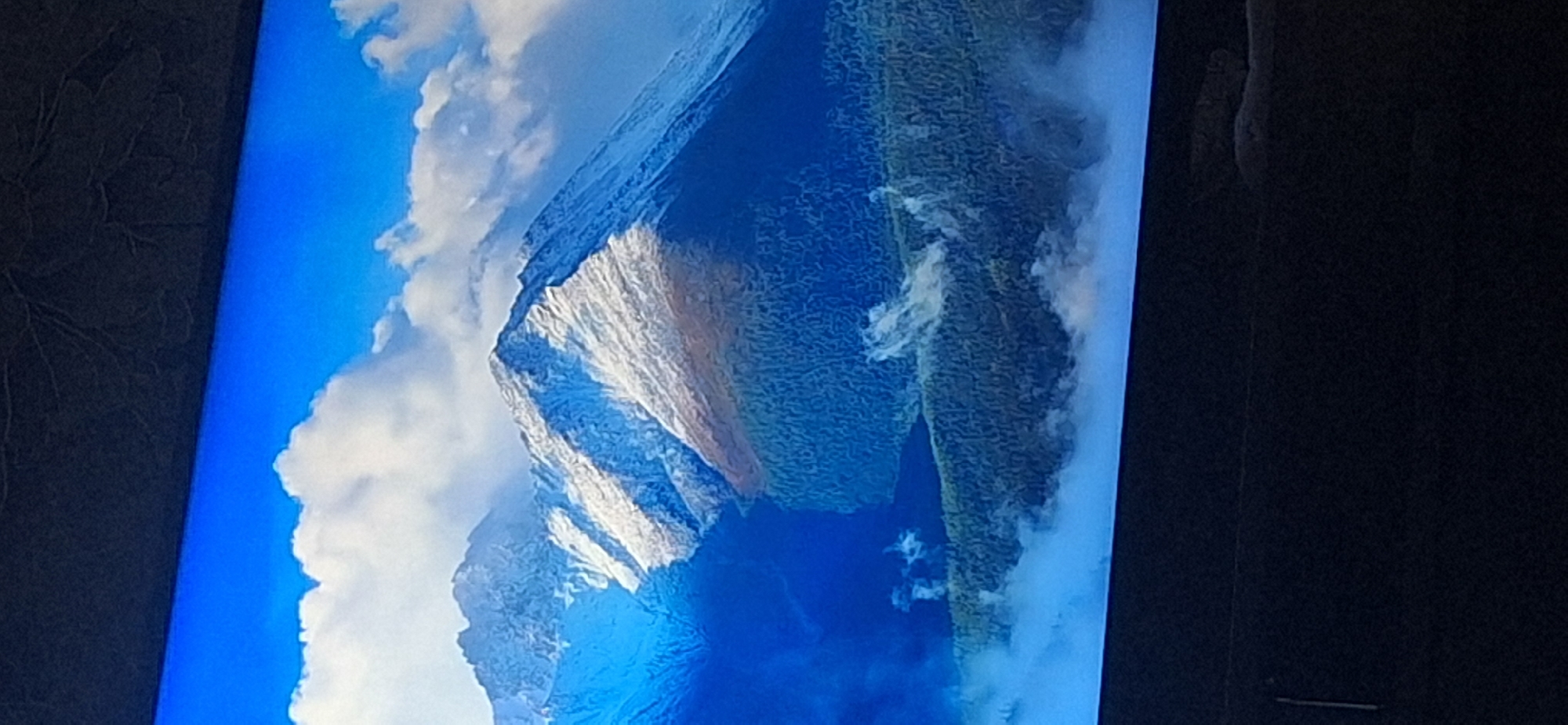 The image displays a television screen displaying a high-angle view of a mountain range. The mountains are snow-capped at their peaks, with darker, forested slopes descending into a misty valley. The sky is a vibrant blue, with scattered white clouds. The image is a photograph of a screen, with the bottom and top edges of the screen appearing black and out of focus. There is a thin vertical line near the bottom left edge of the screen. The image does not depict any specific location in Bila Tserkva, Ukraine.