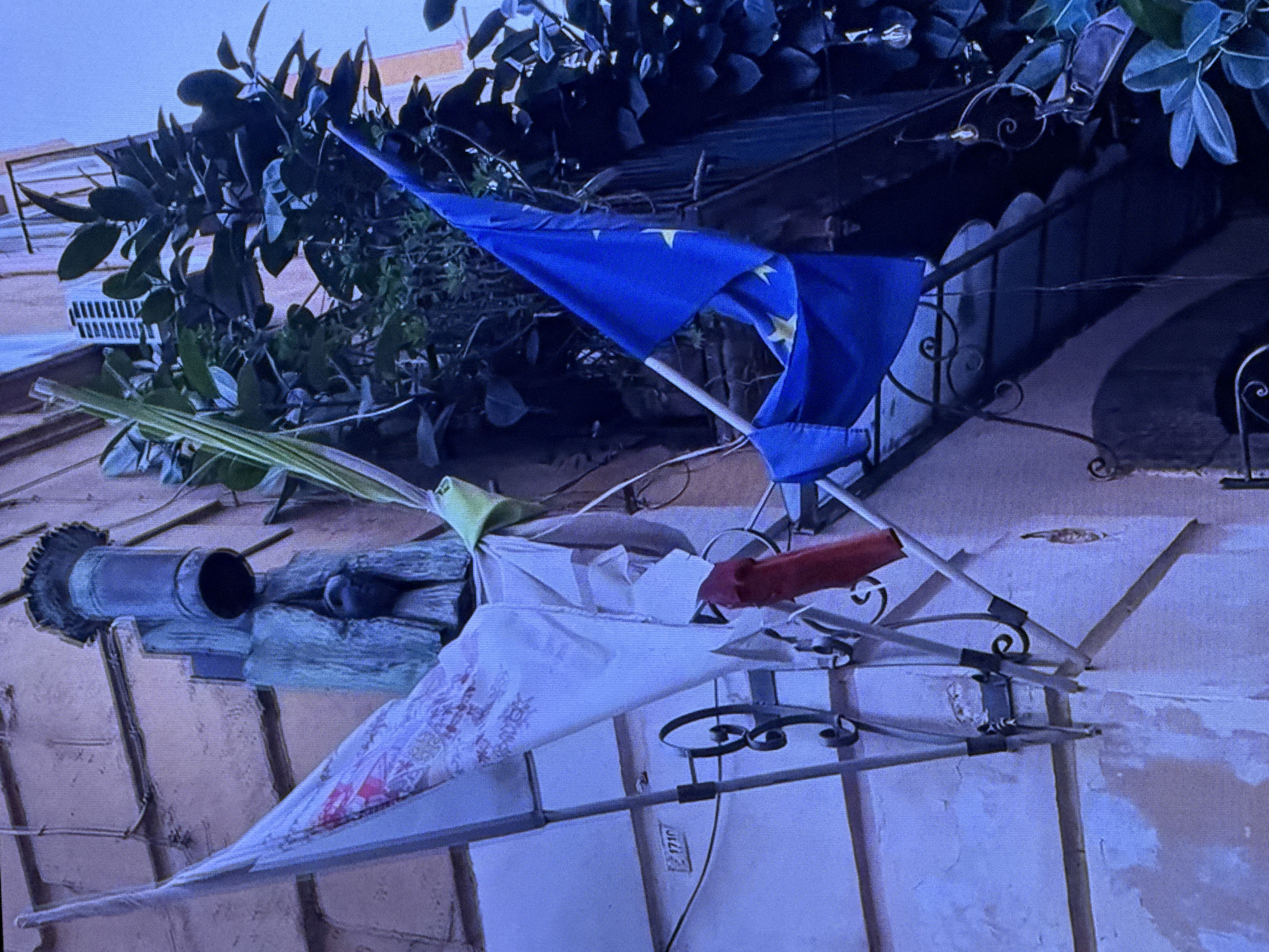 The image shows a close-up, low-angle shot of an outdoor scene with foliage, flags, and a building facade. A blue flag with yellow stars, likely the European Union flag, is prominently displayed, billowing in the breeze. Below it, a white fabric object with a red and patterned design is attached to a metal structure. Green, leafy branches overhang from the top of the frame. A cylindrical object, possibly part of a downspout or architectural feature, is visible on the left. The building facade appears to be made of light-colored, textured material, with some areas showing signs of wear or peeling.
The lighting suggests it could be daytime, possibly with overcast skies or late afternoon light given the subdued colors. The setting appears to be urban, with a building and what might be a balcony or window ledge implied by the visible architecture. The overall impression is a somewhat cluttered yet colorful display, possibly set up for a decorative or symbolic purpose. There is no clear indication of people or a specific event taking place, beyond the static display of flags and fabric. No visible text is discernable in the image.