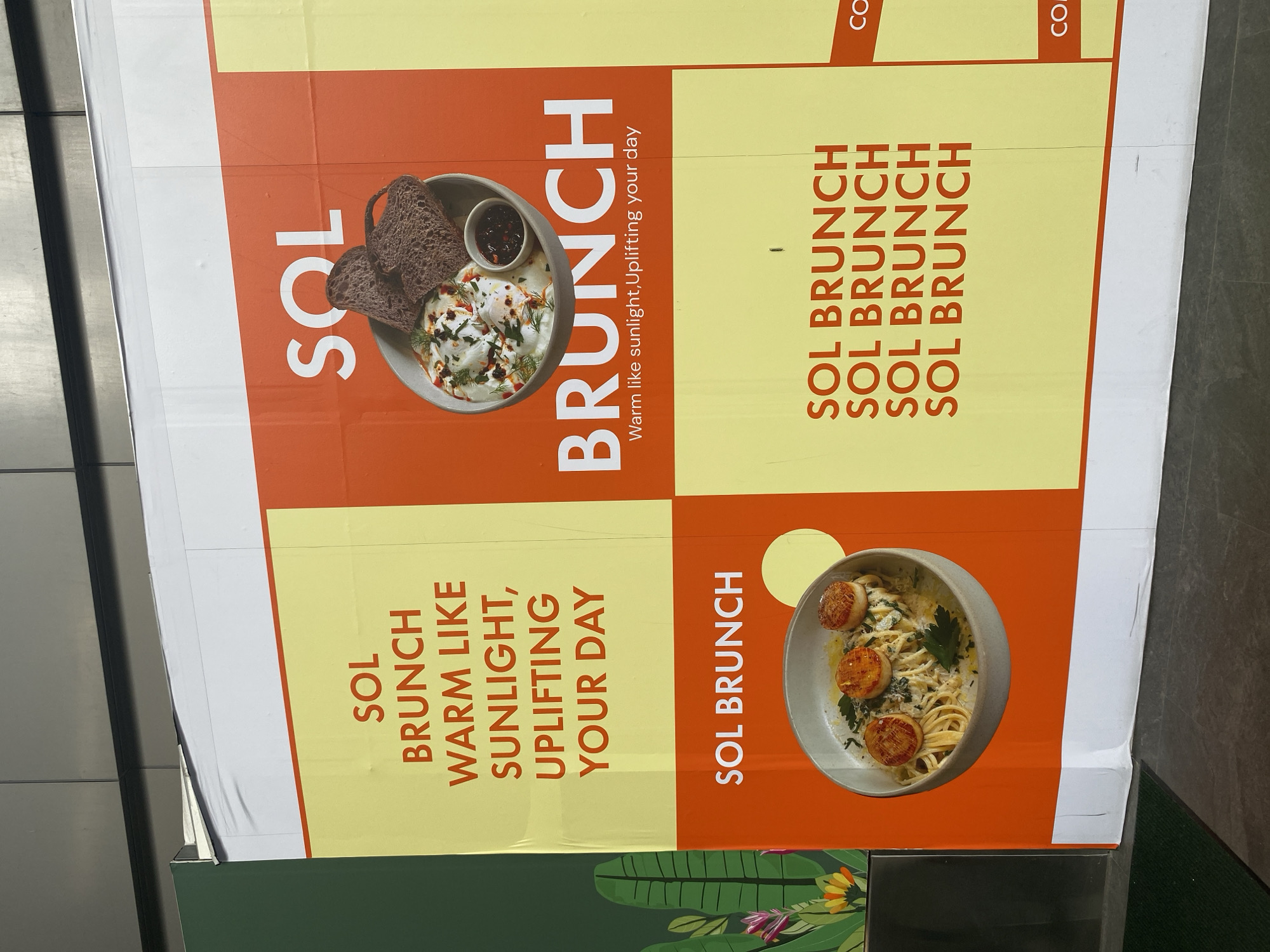 The image shows a large advertisement for "SOL Brunch" with text and images of food. The advertisement is divided into four quadrants. The top left quadrant is orange with white text reading "SOL Brunch" and a photo of eggs, bread, and jam. Below that, the bottom left quadrant is yellow with orange text reading "SOL Brunch Warm like sunlight, uplifting your day." The top right quadrant is yellow with orange text reading "SOL Brunch" repeated four times. Finally, the bottom right quadrant is orange with white text reading "SOL Brunch" and a photo of pasta with scallops. The advertisement appears to be located in Bangkok, Thailand based on the presence of other writing.