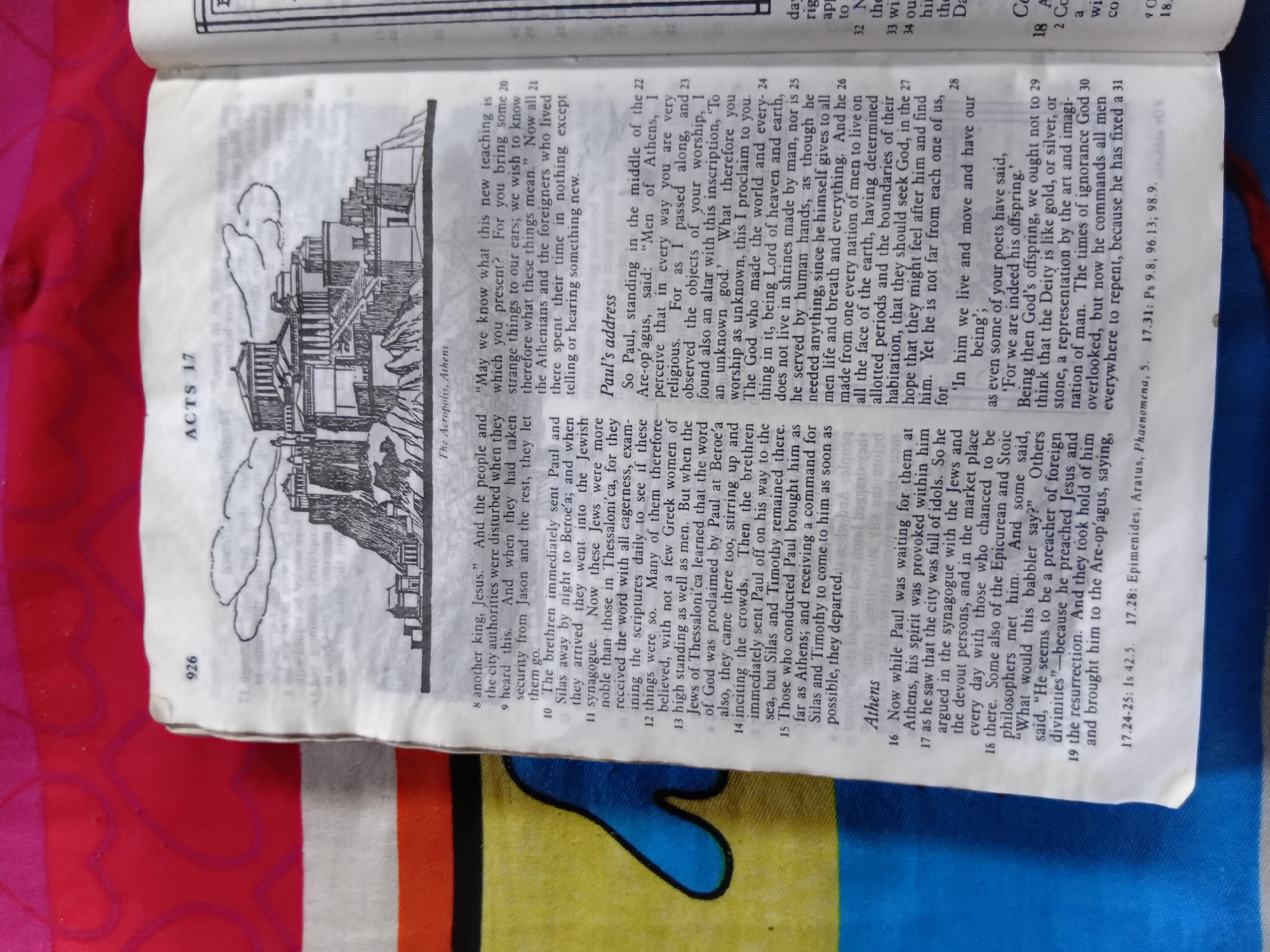 The image shows an open book displaying a passage from the Bible, specifically from the book of Acts, chapter 17. The text discusses Paul's ministry in Athens and his address to the Athenians at the Areopagus.
The left page features a black and white illustration of the Acropolis in Athens, with its prominent ancient structures. Beside the illustration, there is text with a chapter heading "ACTS 17" and the caption "The Acropolis, Athens." The passage itself details events like Paul's interaction with Jews in Thessalonica and Beroea, his journey to Athens, and his encounters with Epicurean and Stoic philosophers.
The right page continues the text of Paul's speech, focusing on his message about the "unknown god" and the nature of the Creator. There are also verse numbers and cross-references to other biblical passages. The text is printed in a dark font on a slightly aged, off-white paper. The book appears to be a study Bible, given the presence of illustrations and annotations. The background of the image, visible beyond the book, is a vibrant, textured fabric in shades of pink and red. The overall impression is of a religious text being studied, with historical and geographical context provided through the illustration and accompanying descriptions.