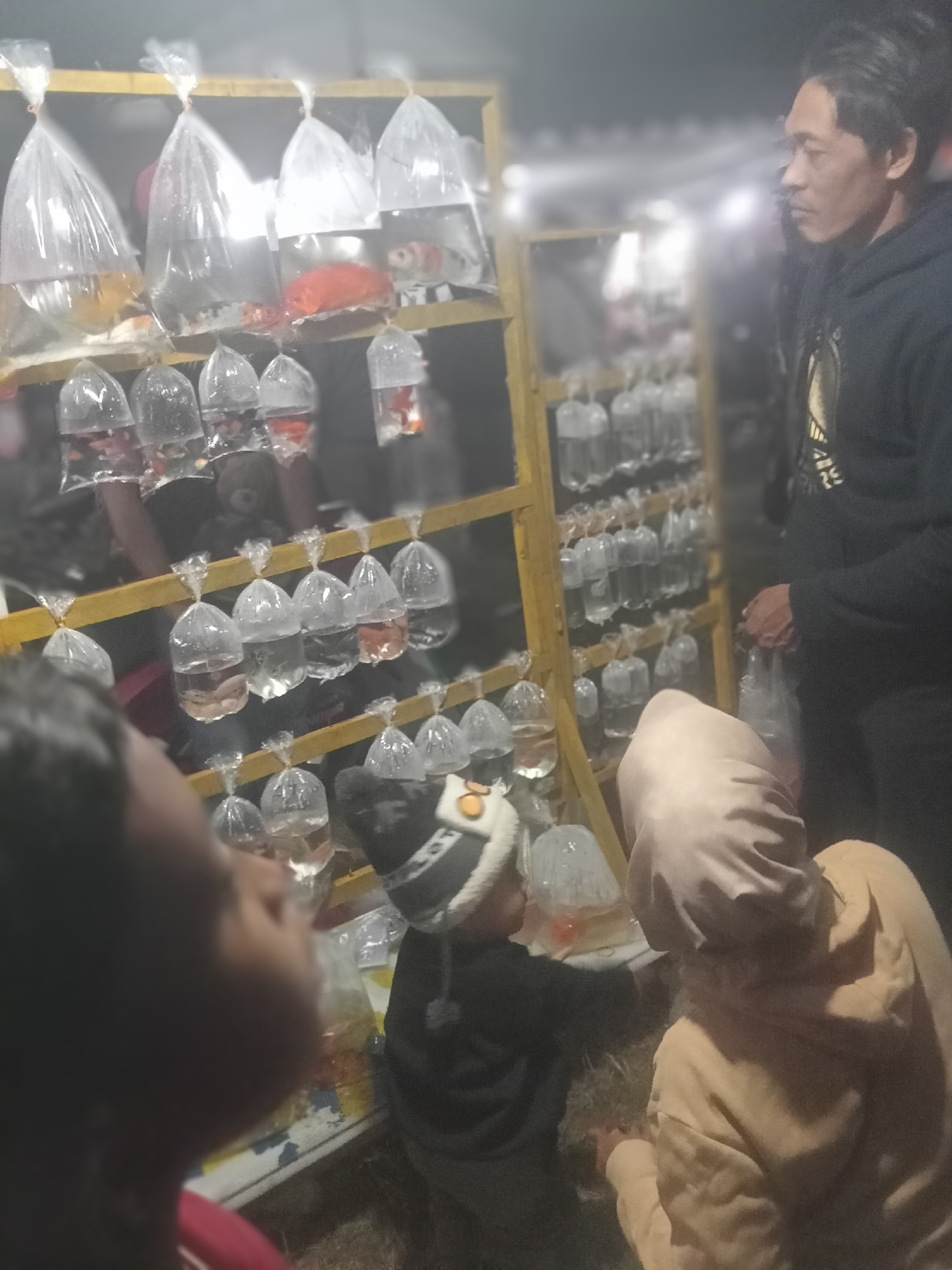 In the city of Pule, Indonesia, a street vendor is selling fish. The vendor, a man, stands near a yellow shelf that holds many plastic bags filled with fish. A child in a hat and a person in a tan hoodie are looking at the fish. The scene is dimly lit, suggesting it might be evening or nighttime. The background is blurred, but it appears to be an outdoor setting, possibly a market.