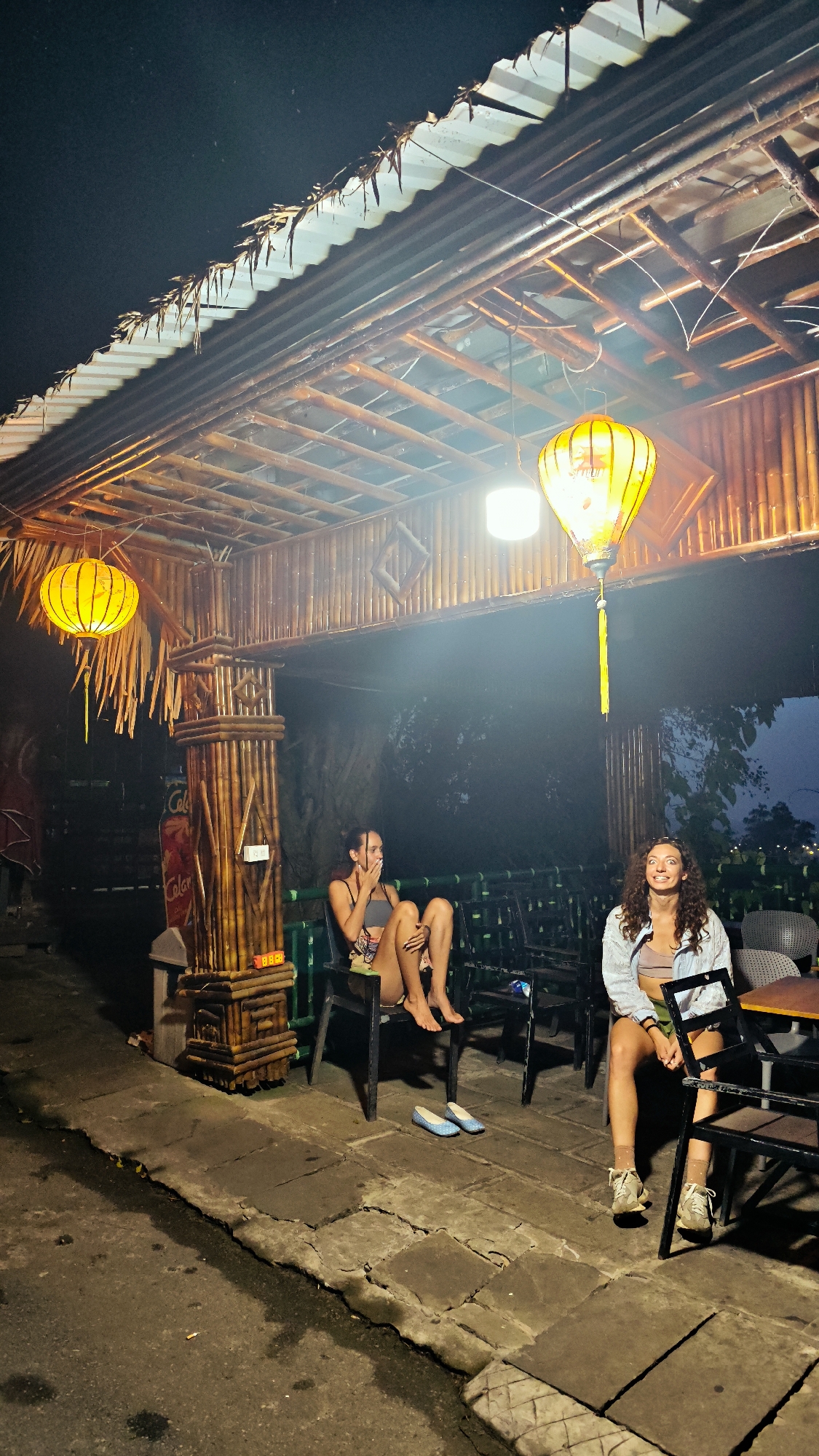 This is a nighttime, eye-level, full shot of two women sitting on chairs under a bamboo structure with a corrugated metal roof. The structure appears to be an outdoor seating area or a covered walkway. The scene is lit by two yellow lanterns hanging from the ceiling of the structure and a white light on the ceiling.
The woman on the left is sitting with her legs crossed and is looking to her left, covering her mouth with her hand. She is wearing a grey tank top and patterned shorts. She has dark hair pulled back. On the floor in front of her are a pair of light blue slip-on shoes.
The woman on the right is sitting with her legs slightly apart and is looking towards the camera with a surprised expression. She is wearing a light-colored button-up shirt over a light-colored tank top and shorts. She has curly, light brown hair.
The structure is supported by thick bamboo poles. The walls of the structure are also made of bamboo. There are several chairs and tables visible in the background, some of which are empty. The ground in front of the seating area is paved with large, irregular stone tiles. The area to the left of the seating area appears to be a street or pathway.
In the background, behind the seating area, there are trees and foliage, and in the far distance, a faint outline of hills or mountains can be seen against the dark sky. The overall atmosphere is relaxed and casual.