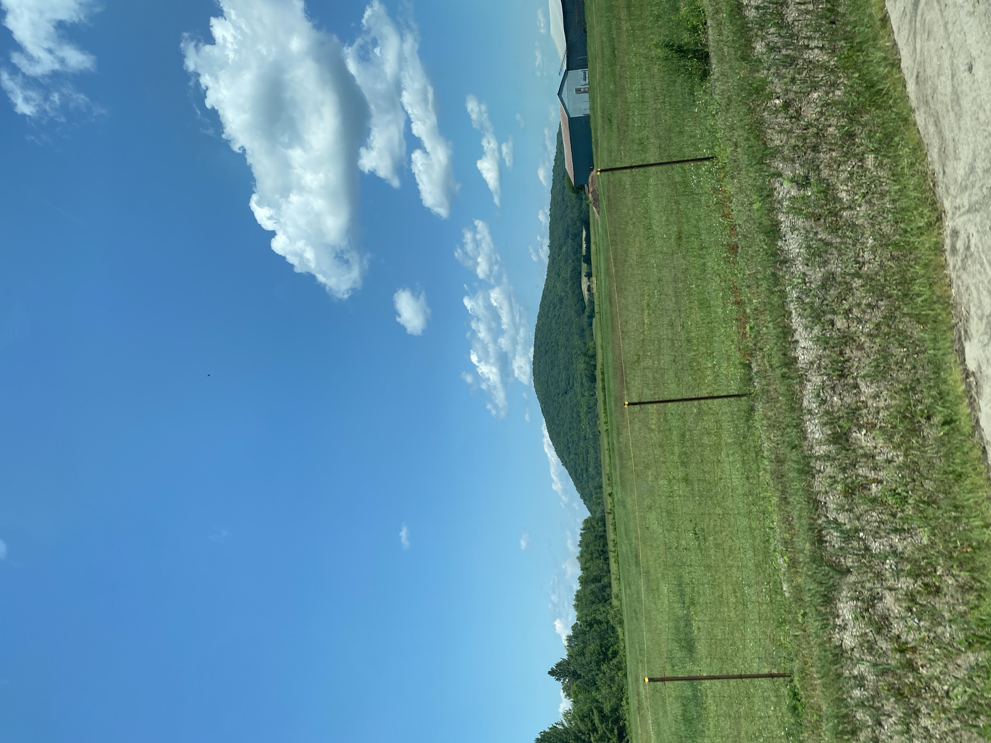 The image captures a serene rural landscape under a bright blue sky dotted with fluffy white clouds. The foreground features a grassy field with a wire fence and two brown posts. Beyond the field, a dense line of green trees forms a natural border. In the distance, a large, verdant hill dominates the horizon. To the right of the hill, two farm buildings with sloped roofs are visible. The overall scene is peaceful and idyllic, suggesting a clear day in a countryside setting. The image does not contain any people or active events.