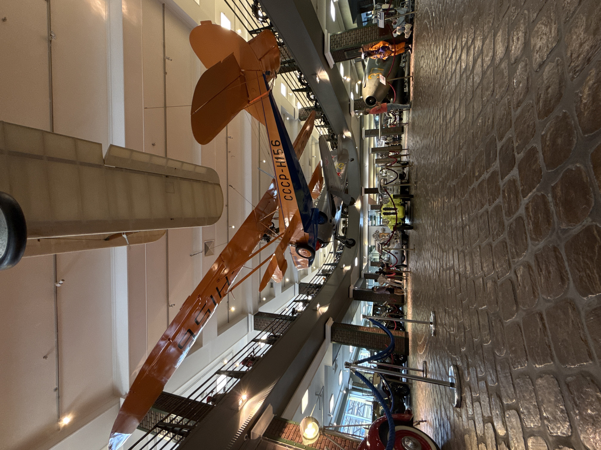 This is an interior photograph of a museum exhibit, likely the Arkhangelskoye Estate Museum in Russia, given the context of the Soviet-era aircraft. The scene depicts a large, multi-level exhibition hall with a brick-patterned floor. Suspended from the ceiling are several vintage aircraft, prominently featuring an orange biplane with the inscription "CCCP-H156" on its wing. Below, on the ground level, various antique vehicles are displayed, including what appears to be a red vintage car on the left, a small white car, and other historical automobiles. A large, olive-green aircraft, possibly a bomber, is positioned on the right, with a figure in an orange flight suit standing beside it. People are visible on both the ground floor and on the upper balconies, observing the exhibits. The environment is well-lit, with visible light fixtures on the ceiling and spotlights illuminating some of the displays. The architecture features brick pillars and metal railings on the upper levels.