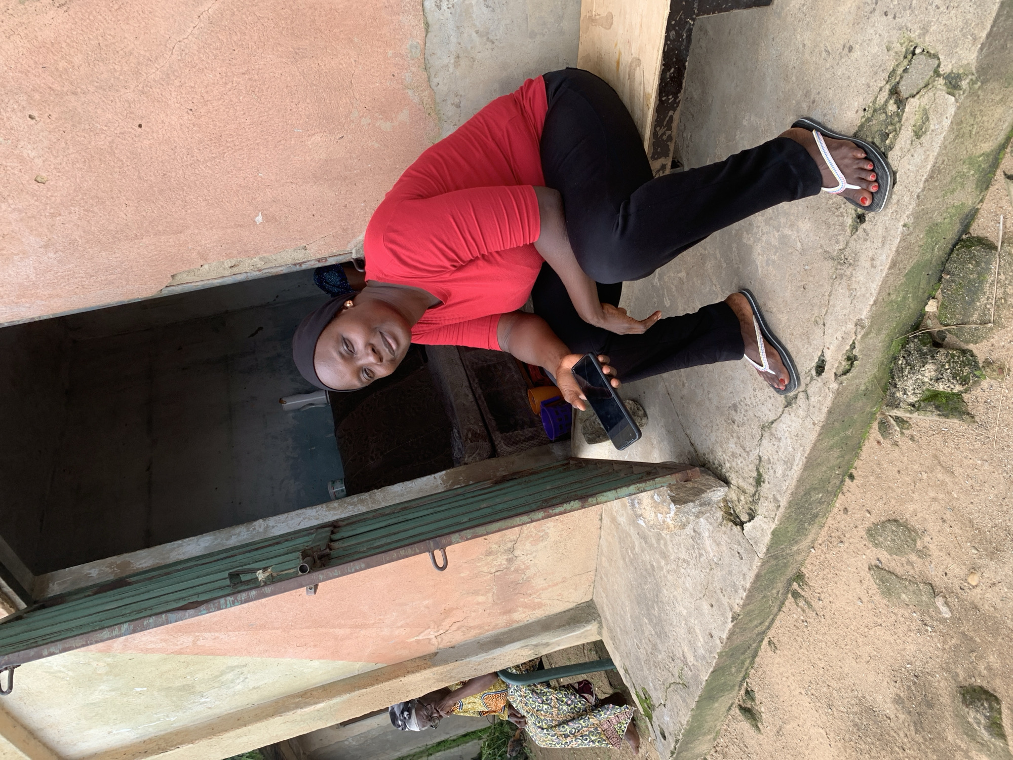 In Sabon Tasha, Nigeria, a woman wearing a red shirt and black pants is sitting on a wooden bench outside a building. She is holding a black smartphone in her right hand and looking towards the camera with a slight smile. Her feet are adorned with black flip-flops with white straps. To her left, an older woman in a colorful patterned dress and headscarf is seated on a metal chair, facing away from the camera. The building behind them has a peach-colored wall and a green metal folding door that is partially open, revealing a dark interior with what appears to be stored items. The ground in front of the building is a mixture of concrete and sandy soil, with some scattered rocks and moss.