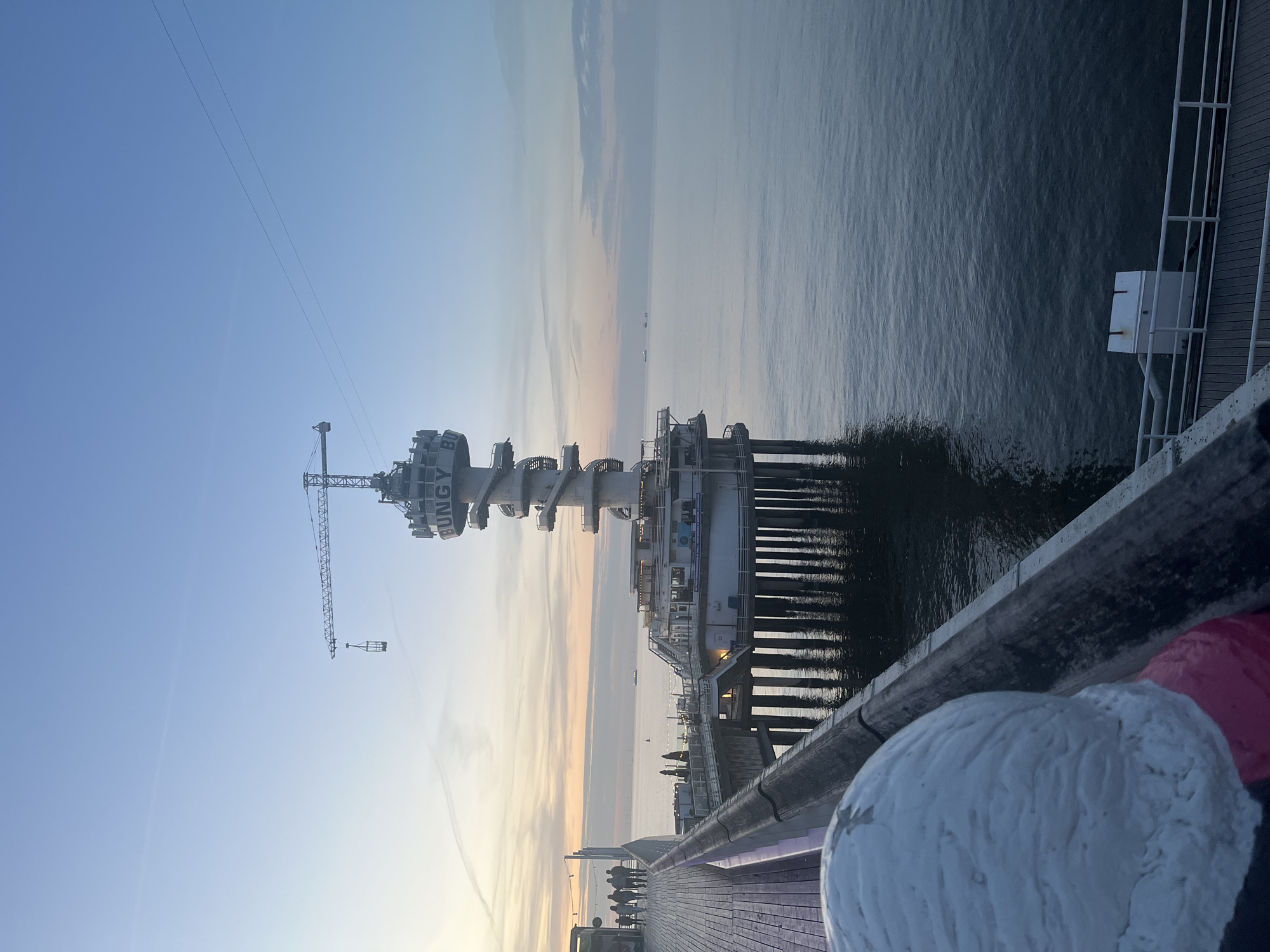 The image depicts a view from a pier in The Hague, Netherlands, at dusk or dawn. A prominent structure, the Pier Scheveningen, stands tall, featuring a tower-like structure with a spiral staircase and a crane attached. The sea stretches out into the horizon, with a few distant ships visible. The pier's wooden deck is in the foreground, with a railing separating it from the water. A bollard is visible, partially obscured. Several people are walking and standing on the pier. The sky is a mix of blue and orange hues, indicating sunrise or sunset. The water reflects the sky's colors.