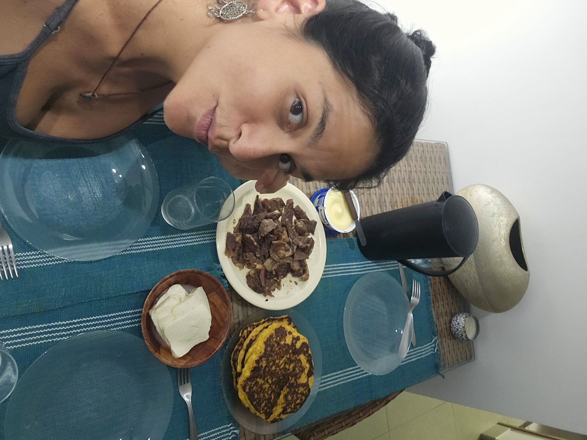 A person is positioned next to a table setting. The setting includes two clear plates, a glass of water near each plate, and forks set out for each place setting. On the table, there are also plates containing food items: one plate holds a portion of beef, another contains what appears to be cheese in a wooden bowl, and a third plate is piled with pancakes. A black object is also on the table. The table is covered with a blue and white striped tablecloth. The location of the scene is unknown, in the city of Unknown, Unknown.