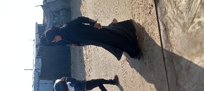 A person in a black hijab and dress stands on a dirt ground next to a building with a banner. Another person wearing a jacket and pants stands below the first person. Shadows suggest the photo was taken on a sunny day.