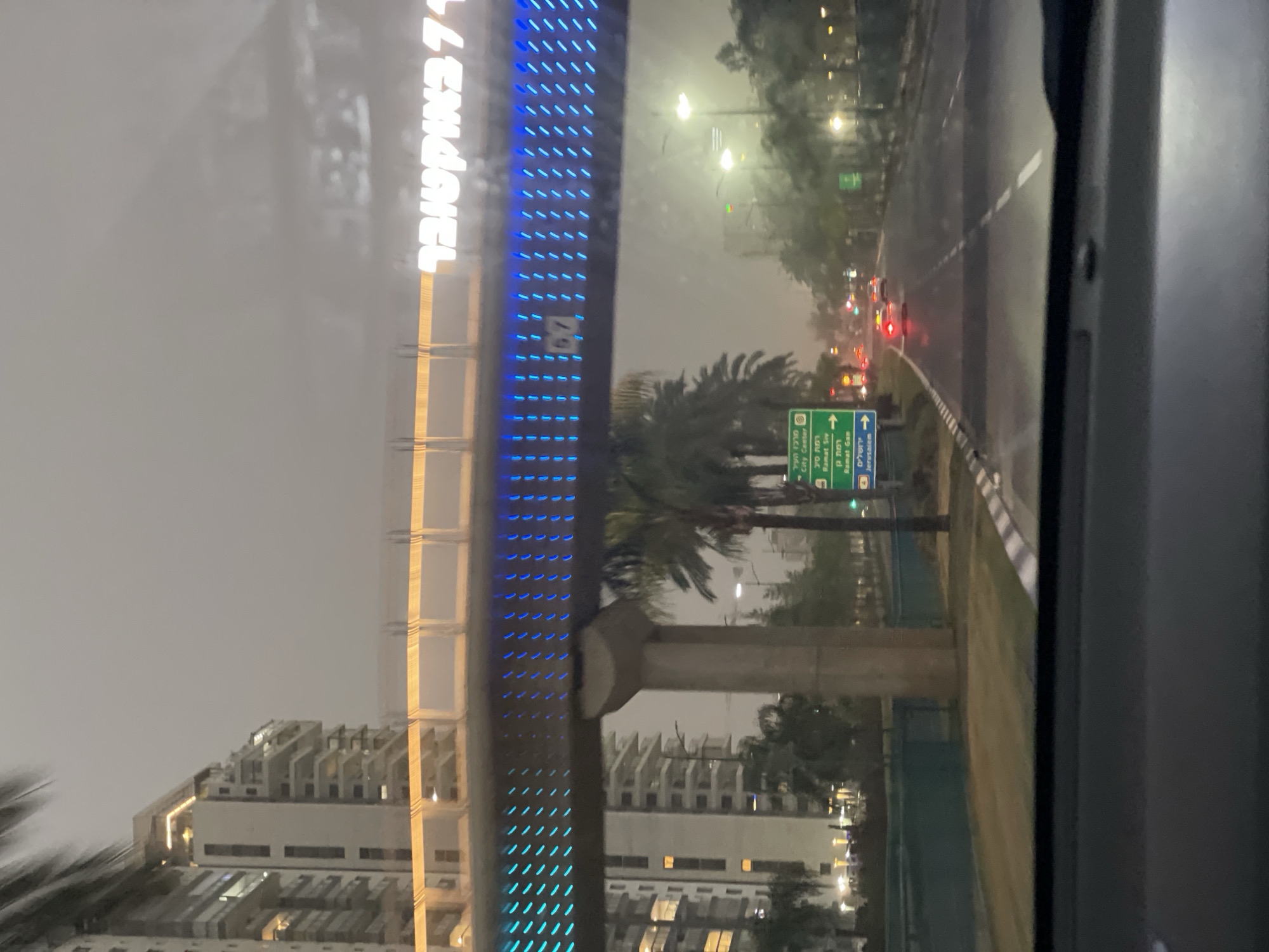The image shows a street scene in Petah Tikva, Israel, on a rainy day. The view is likely taken from inside a vehicle, possibly a car or bus, as part of the vehicle frame is visible. A large illuminated sign, possibly advertising or marking a location, reads "71 באוקטובר" (71 October). Street signs are visible, one indicating directions to the "City Center", "Ramat Siv", "Ramat Gan", and "Jerusalem". Elevated construction is present, possibly related to a transportation system or building project. In the background, buildings are visible, along with streetlights and other vehicles on the road. The atmosphere is overcast and wet, with reflections on the road surface.