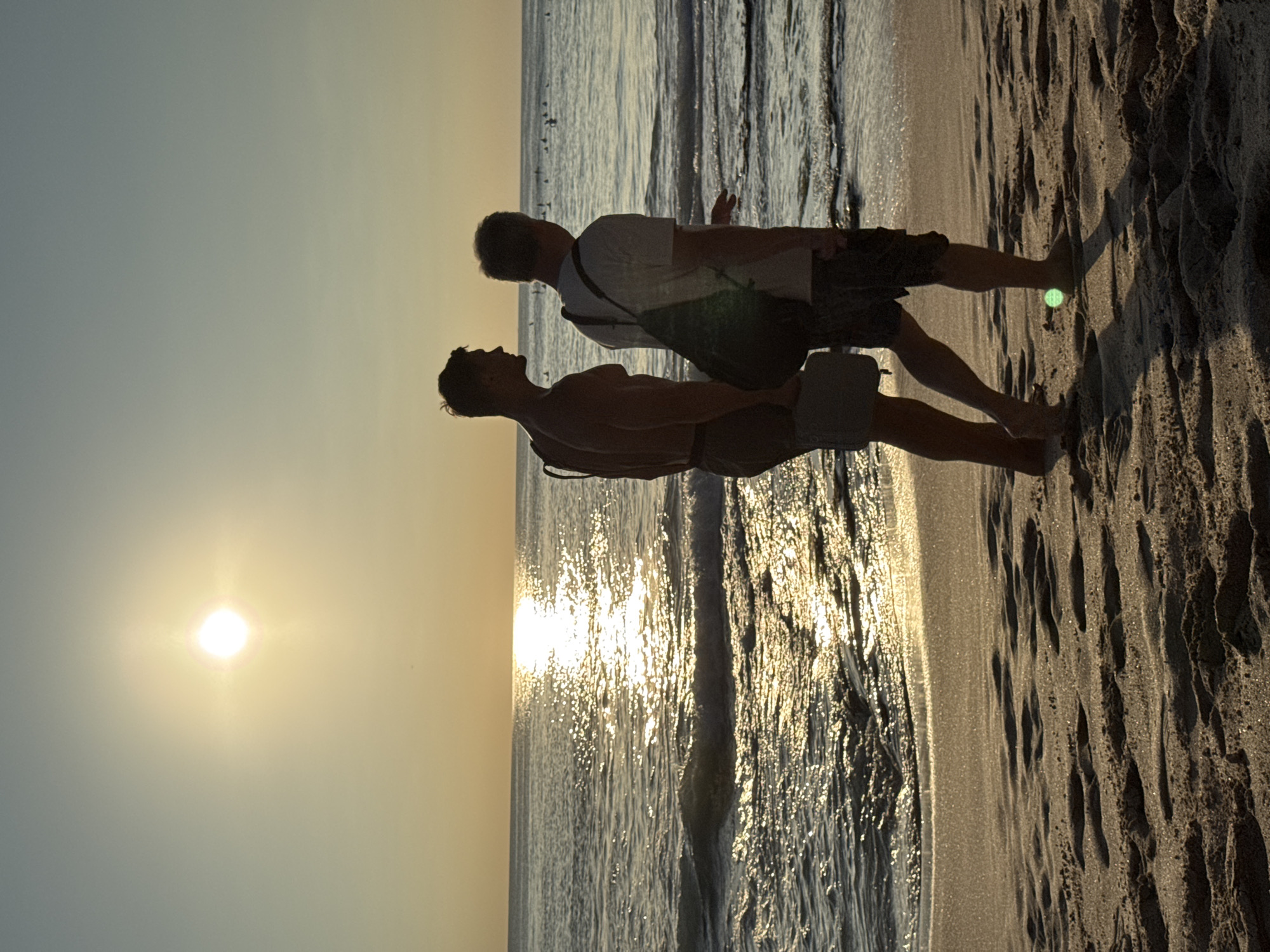 Two males stand on the beach. One wears a white shirt and dark shorts with a strap or bag. The other male has bare skin from the chest up and wears light-colored shorts. The beach is wet from the tide. The sun is setting in the distance, reflecting off the water. The location is Kuta Selatan, Indonesia.