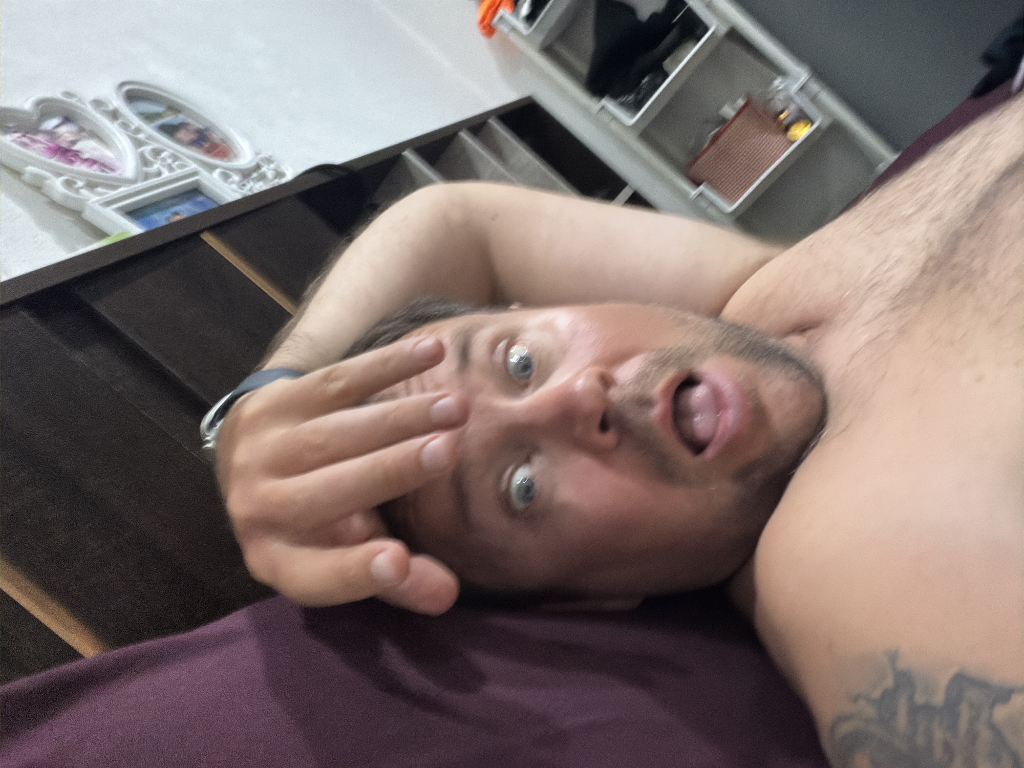 A man is lying on a bed in Baan Mae Nam, Thailand, with his hand raised to his forehead, his eyes wide and mouth open. His body is bare-chested and he has tattoos on his left arm. The room appears to be a bedroom, with a dark headboard and a dresser with a photo frame containing multiple pictures. A shelf unit is visible in the background, with various items stored on it.