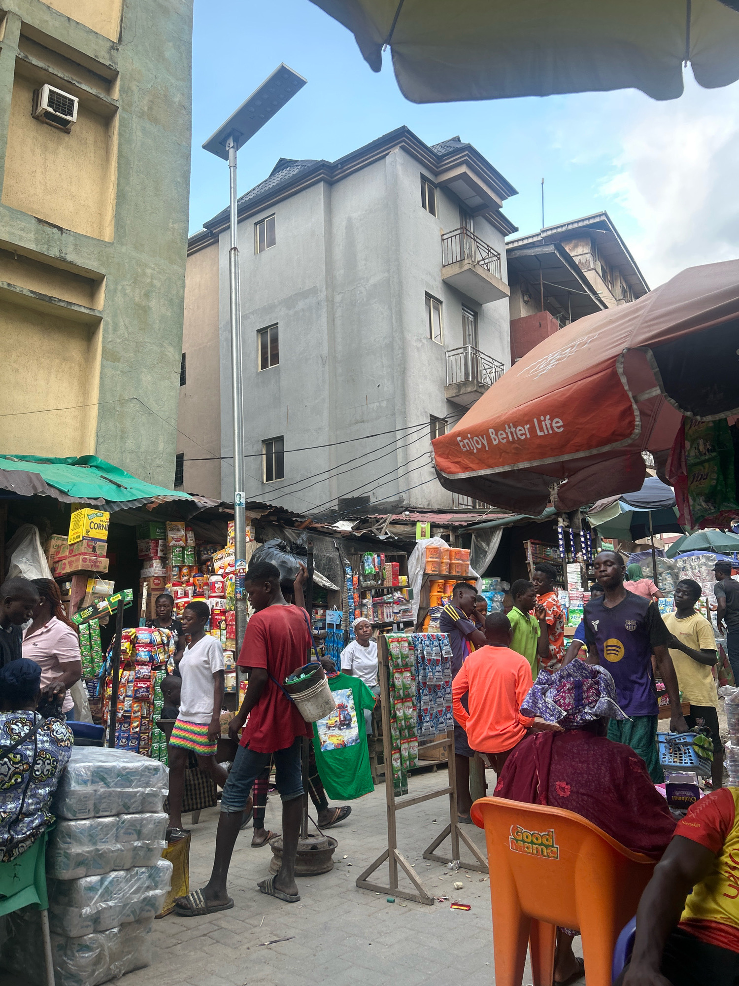 The image depicts a bustling street market scene in Lagos, Nigeria. A narrow street is lined with stalls selling various goods, including packaged food items, drinks, and clothing. Several people are present, engaged in shopping, socializing, and carrying out their daily activities. The architecture suggests an urban environment, with multi-story buildings visible in the background. The overall impression is one of lively commerce and everyday life in a Nigerian city.