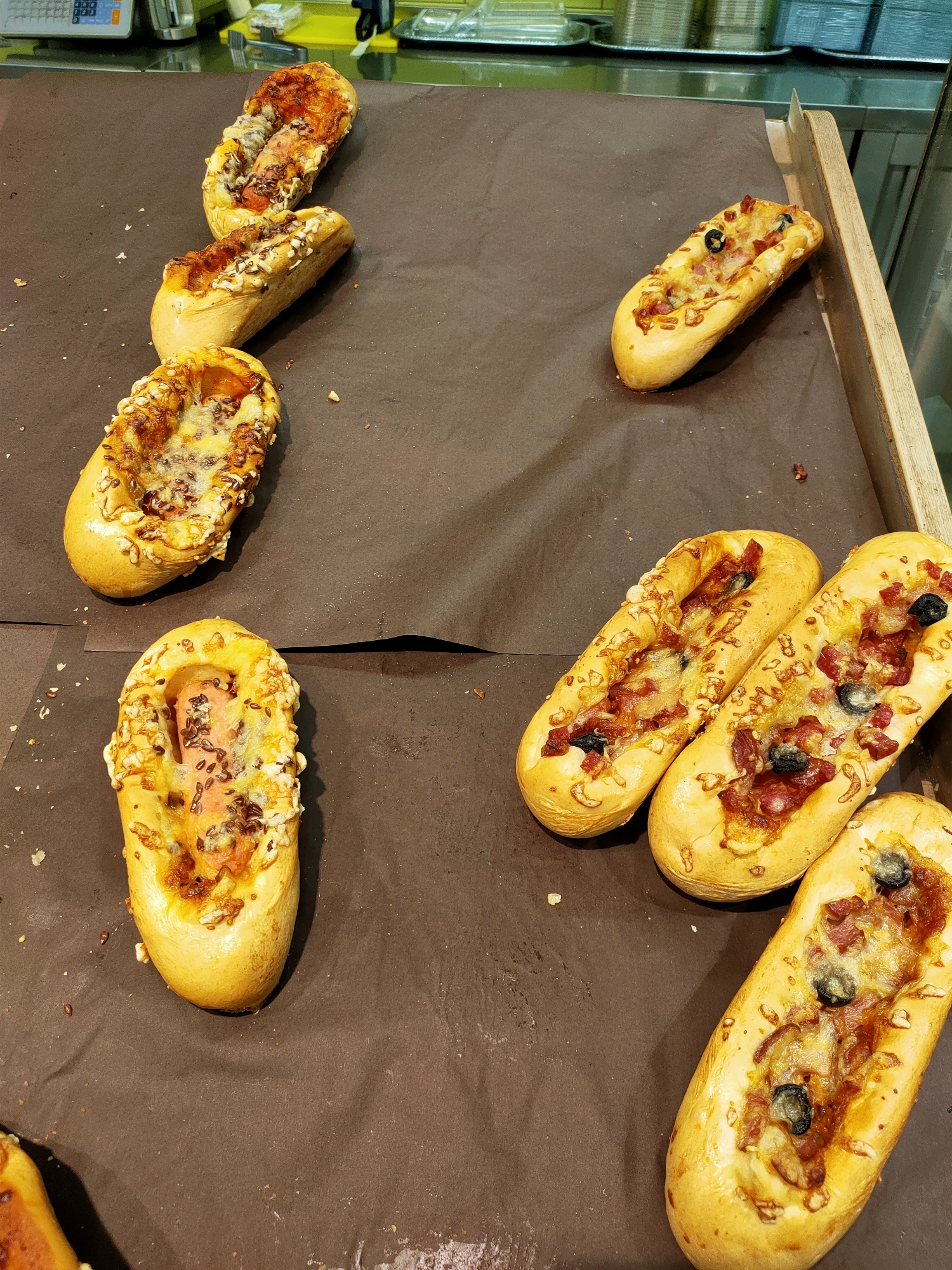 This is a top-down, eye-level shot of several baked goods, likely pizzas or savory pastries, displayed on a dark brown surface. The pastries are elongated and appear to be filled with various ingredients, including melted cheese, what looks like sausage or pepperoni, and some have visible olives or seeds. The lighting suggests an indoor setting, possibly a bakery or a food stall. The background shows a metal counter and some yellow shelving, which might indicate a commercial kitchen or a retail environment. There are no people visible in the image. The pastries are arranged in a casual, somewhat scattered manner, suggesting they are ready for sale or display.